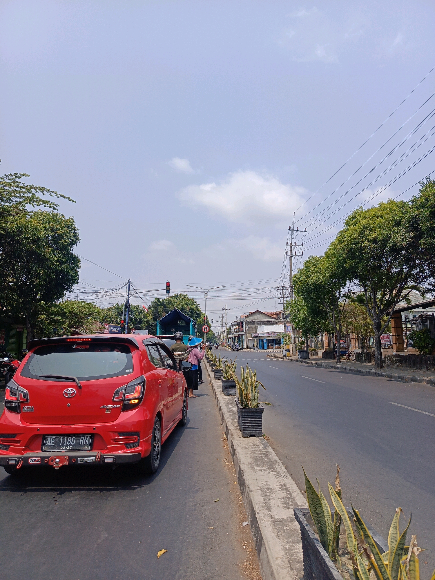 The image depicts a street scene in Sukowinangun, Indonesia, under a bright, partly cloudy sky. In the foreground, a red car is parked on the side of the road, facing away from the viewer. The car has a license plate that reads "AE 1180 RM" and a sticker for "02-27". To the right of the car, a person in a pink hijab and dark clothing stands near a blue tarp-covered vehicle. Further down the road, there are buildings on the left and right sides, with trees lining the street. Traffic lights are visible, indicating an intersection. The road is paved, and a sidewalk runs along the right side of the image, adorned with potted snake plants. Electrical wires crisscross the sky, supported by utility poles. The overall scene suggests a typical day in a bustling Indonesian town.
