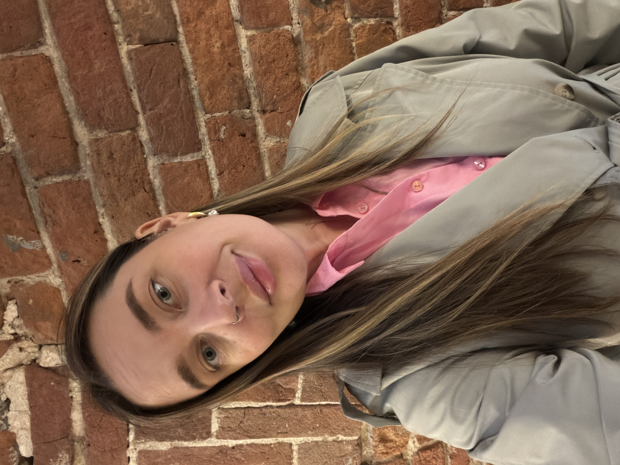 A young woman is looking at the camera. She is wearing a pink button-down shirt and a light grey jacket. She has brown hair and light eyes, and a nose ring. The background is a brick wall. There is no information to suggest that the location is Moscow, Russia.
