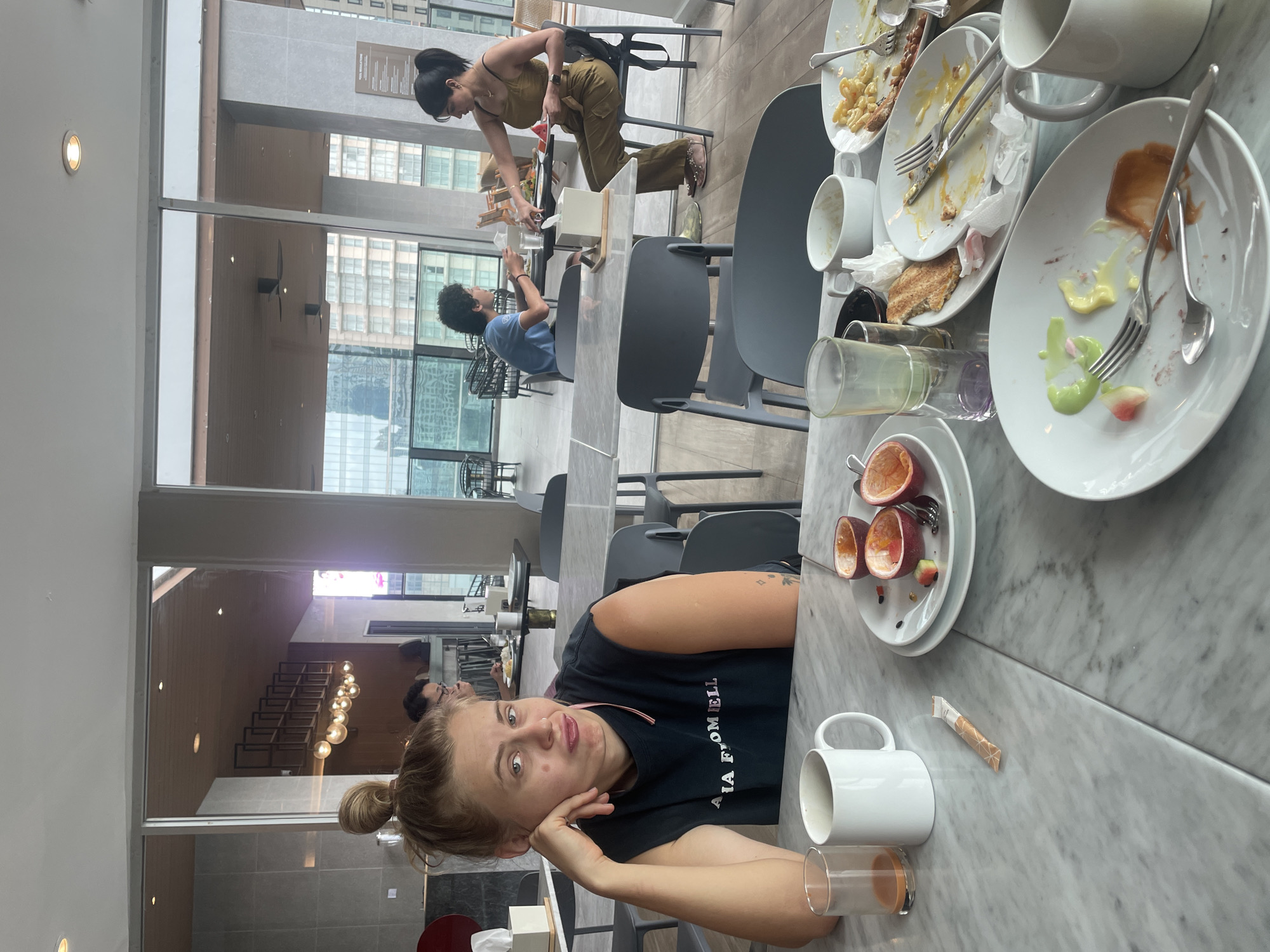 The image depicts an indoor dining scene, likely a breakfast or brunch, in a modern restaurant or hotel in Bangkok, Thailand, during the daytime.
In the foreground, a young woman with blonde hair tied in a bun is looking directly at the camera with a playful, pouting expression, her left hand resting on her chin. She is wearing a black t-shirt with the text "VIA FROMNELL" partially visible. The marble-patterned table in front of her is strewn with used white plates containing food remnants, including macaroni, toast, and colorful purees. Forks, spoons, white mugs, clear glasses (one with a drink, another empty), and a plate with two halves of a passion fruit are also visible. A small, wrapped packet lies on the table.
Beyond the foreground, the spacious dining area features large glass windows, through which modern city buildings with numerous windows are visible. Reflections on these windows create layered imagery, showing interior elements such as a person (likely male with dark curly hair) seated at a table, appearing to look at a phone. Further into the room, another woman with dark hair, dressed in an olive-colored top and pants, is standing and leaning over a table. Another individual in a blue t-shirt is also seated further back.
The setting is contemporary, with dark gray chairs and light-colored walls. A sign on a distant wall faintly reads "NEW RESIDENCE" and possibly "HOTEL," confirming the type of establishment. The scene conveys a relaxed and informal atmosphere following a meal.