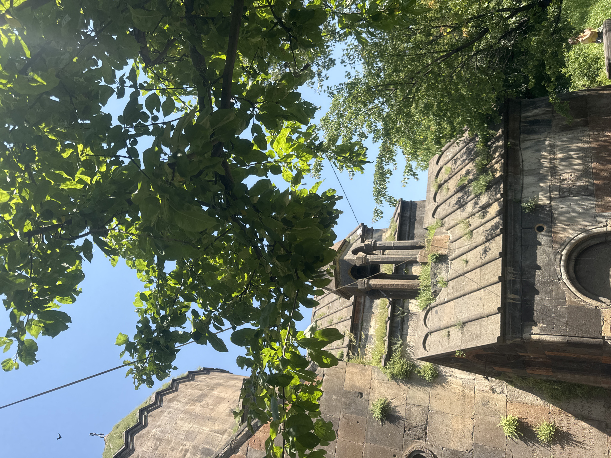 In Bjni, Armenia, a stone bell tower stands next to an ancient stone building with a domed roof. Green leaves from a nearby tree overhang the scene, casting shadows on the stone structures. The sky is clear and blue. The architecture suggests a historical or religious site, likely a church or monastery. A small bird is visible in the sky near a distant, smaller structure. A person is visible in the lower right corner of the image, sitting on a stone ledge.
