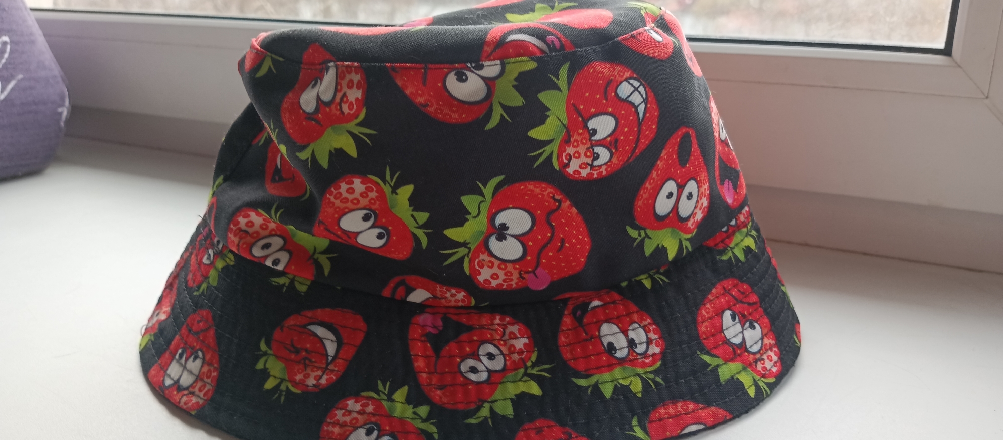 The image features a close-up of a bucket hat adorned with a playful pattern of cartoon strawberries. The hat's fabric is black, serving as a dark background for the vibrant red strawberries, which also have green leafy stems. Each strawberry is anthropomorphized with white eyes and black pupils, displaying a variety of human-like expressions, including smiles, wide-eyed surprise, and some even sticking out their tongues. The hat is positioned on a white windowsill, with a faint shadow cast beneath it. In the background, a white window frame is visible, and beyond the window, an out-of-focus outdoor scene, likely a dull or overcast sky with hints of trees, can be discerned. To the far left, a blurry, purple-colored object with some white markings is partially visible. The scene is illuminated by natural daylight coming from the window. There is no discernible activity taking place, and no visible text on the hat or in the surrounding environment.