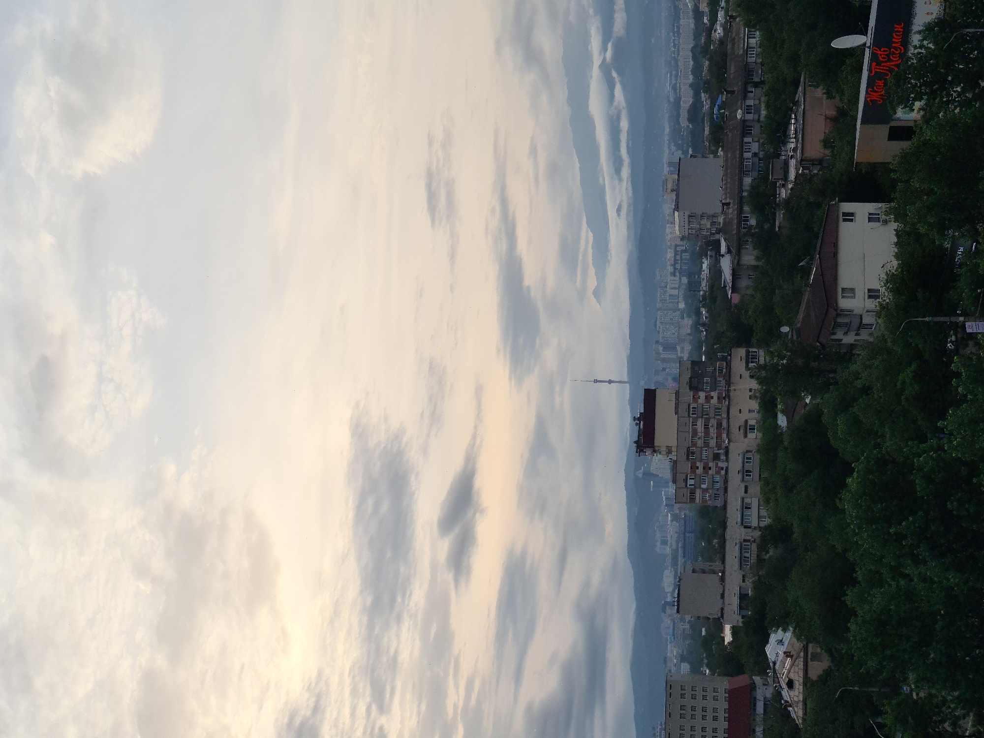 The image captures a panoramic view of Almaty, Kazakhstan, under a cloudy sky. In the foreground, dense green trees form a canopy over residential buildings. Beyond the trees, mid-rise apartment buildings, characteristic of Soviet-era architecture, are visible. Further in the distance, a cityscape unfolds with taller modern buildings and the iconic Almaty Television Tower stands on a hill. The background is dominated by the majestic Tian Shan mountains, partially obscured by clouds and mist, suggesting a mild or humid day. The overall impression is one of a sprawling urban environment nestled against a dramatic natural landscape.