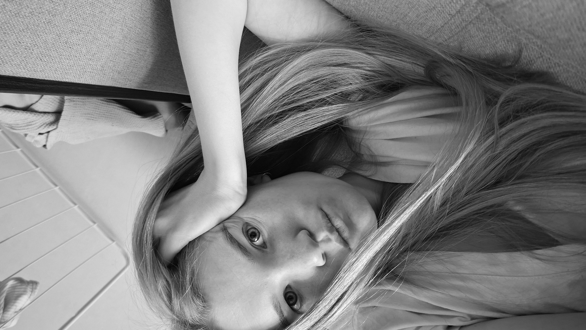 A monochrome portrait depicts a young person, likely female, with long, light-colored hair, looking directly into the camera. The image is oriented sideways, with the subject's head positioned towards the upper right if rotated to a standard upright view. The subject's face is central, characterized by wide, direct eyes and slightly parted lips, conveying a contemplative or introspective expression.
Long, flowing hair, appearing blonde or light brown in black and white, cascades around the head and shoulders, spreading across a light-colored, soft garment. One arm is bent, with the hand resting near the side of the head, partially framing the face. The other arm is also partially visible. A small earring or ear accessory is faintly visible on one earlobe.
The setting appears to be an indoor environment, possibly a living room or bedroom. In the upper left background, a textured, fabric surface, resembling upholstery from a sofa or bed headboard, is visible. The lower left background shows a lighter, possibly tiled or slatted surface, hinting at a floor or a piece of domestic furniture like a drying rack. The lighting is soft and even, emphasizing facial features and hair texture. The black and white tonality lends a timeless and intimate quality to the image. There are no specific visual cues within the image to indicate the time of day, weather, or a precise geographical location. No visible text is present.