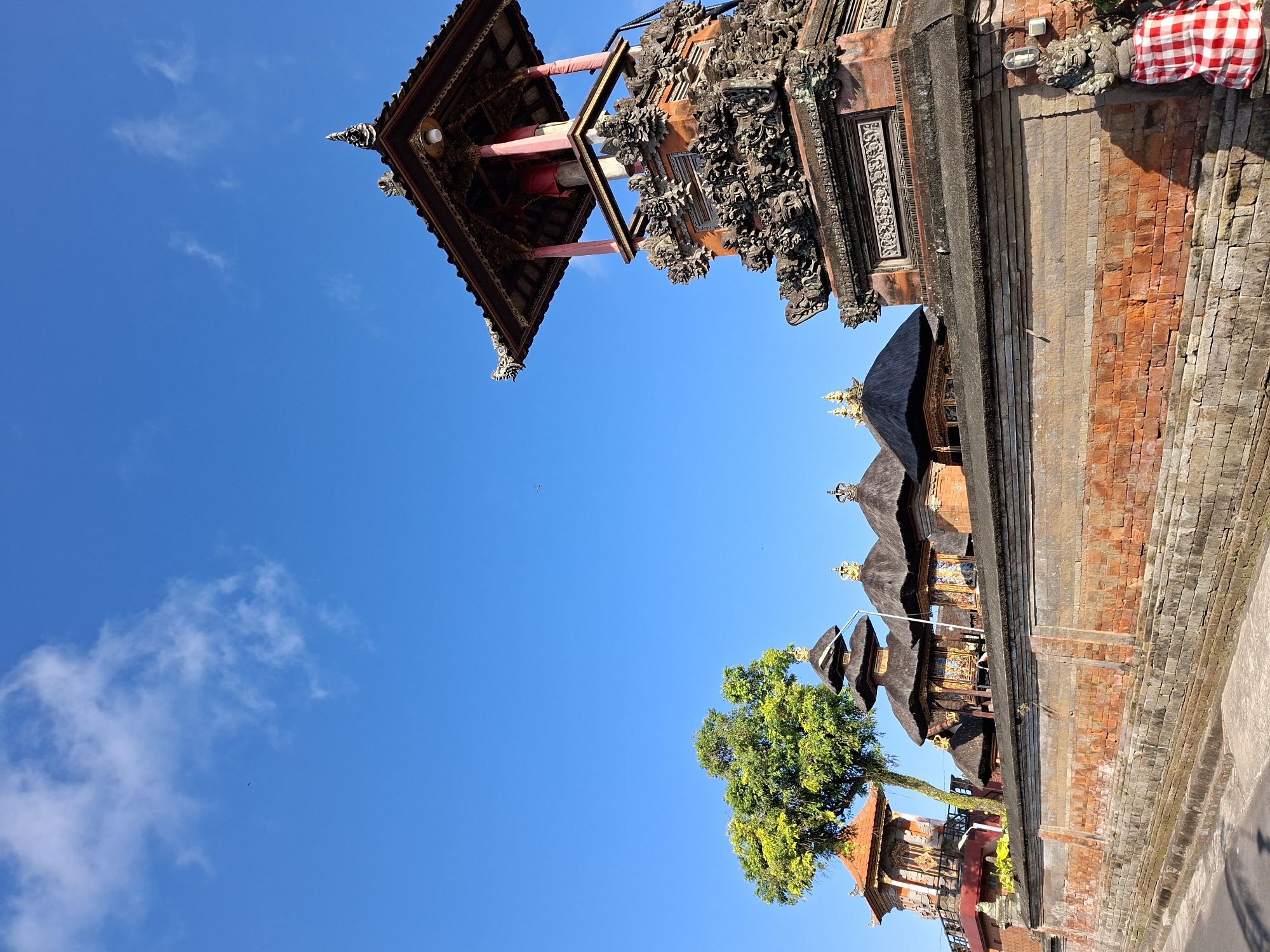 The image shows a view of the Pura Mangkunegaran temple complex in Blahbatuh, Bali, Indonesia. The foreground is dominated by a weathered brick wall, some sections of which are covered in moss. To the right of the wall, a stone statue of a guardian figure, adorned with a red and white checkered cloth, stands near a modern outdoor light fixture. Behind the wall, several traditional Balinese structures with tiered roofs are visible. One prominent structure is a tall, ornate tower with intricate carvings and a wooden roof. A large, leafy green tree stands to the left of these buildings. The sky above is a clear, vibrant blue with a few wispy white clouds. The overall scene depicts a cultural and architectural landmark under a bright, sunny sky.