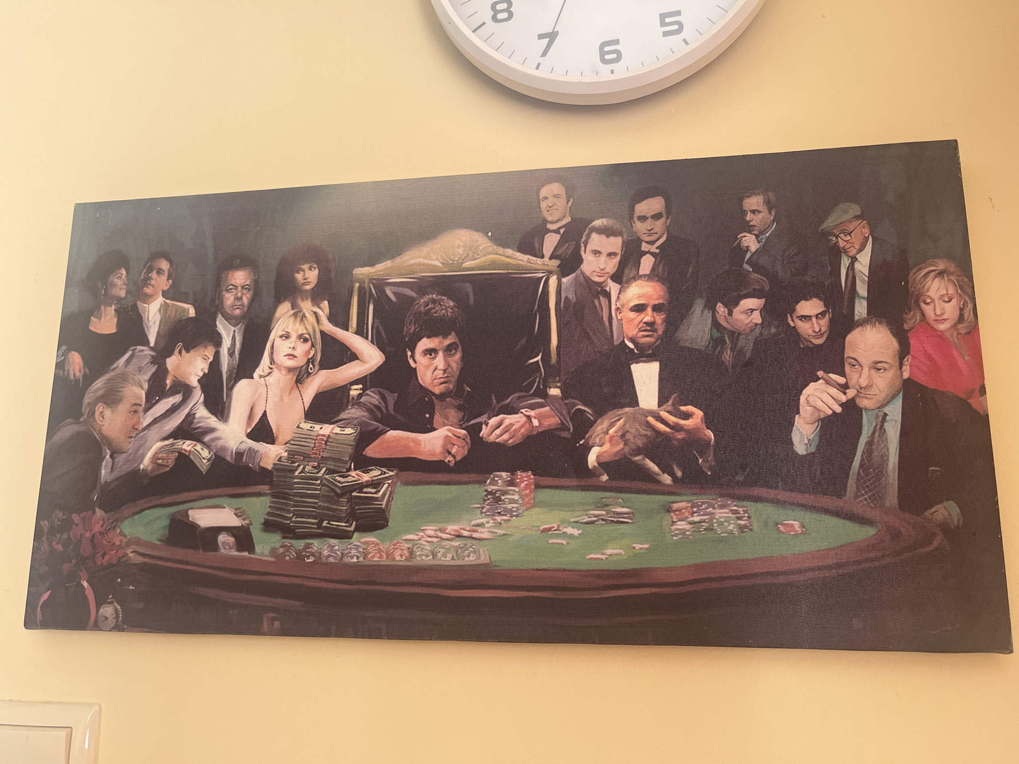 A photograph captures a large, rectangular print or painting affixed to a light yellow wall in the city of Burgas, Bulgaria. A white circular wall clock, displaying black numerals and hands, is partially visible in the upper right quadrant of the photographic frame, positioned above the artwork.
The artwork itself depicts a group of approximately 20 individuals, predominantly male, engaged in what appears to be a poker game. The figures are recognizable characters from various gangster and crime films, including Tony Montana, Michael Corleone, Vito Corleone (both young and old), and Tony Soprano. They are arranged around a large, oval-shaped poker table with a green felt surface and dark wood trim.
On the table, numerous stacks of U.S. dollar bills are visible, particularly on the left side, along with scattered poker chips in various colors (red, blue, purple, white). A dark vase containing red flowers and a small metallic timer-like object are situated on the far left edge of the table.
Key figures include:
- A blonde woman in a black strappy top, positioned left of center, with large stacks of cash near her.
- Tony Montana (Al Pacino) in a dark shirt, seated center, holding a cigar.
- Michael Corleone (Al Pacino) in a dark suit and bow tie, seated to the right of Tony Montana, holding a cat in his lap.
- Tony Soprano (James Gandolfini) in a suit on the far right, holding a cigar.
- Young Vito Corleone (Robert De Niro) in a suit, visible on the far left.
- Older Vito Corleone (Marlon Brando) in a suit and bow tie, seated center-right.
The individuals are depicted in various states of attention, some looking at cards, others observing the scene, and some directly at the viewer. The overall ambiance is dark and atmospheric, consistent with the thematic portrayal of the characters.