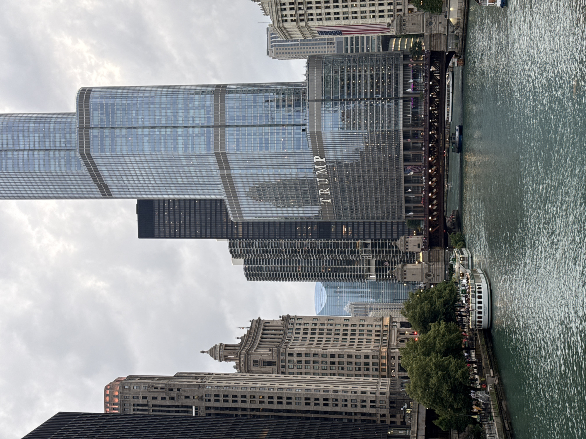 This image captures a view of the Chicago River in Chicago, Illinois, United States of America. The scene is dominated by several prominent skyscrapers, including the Trump International Hotel & Tower, which is the tallest building in the frame and features the word "TRUMP" in large lettering on its facade. To the left of the Trump Tower are other skyscrapers, including the historic Wrigley Building and other commercial buildings. The Chicago River flows in the foreground, with several tour boats visible on its surface. One boat, labeled "EMERALD LADY," is docked along the riverwalk, and several other boats are in motion. The riverbanks are lined with trees and walkways, where pedestrians can be seen. A bridge, likely the Michigan Avenue Bridge, spans the river in the mid-ground, with traffic visible on its surface. The sky is overcast, suggesting a cloudy day.