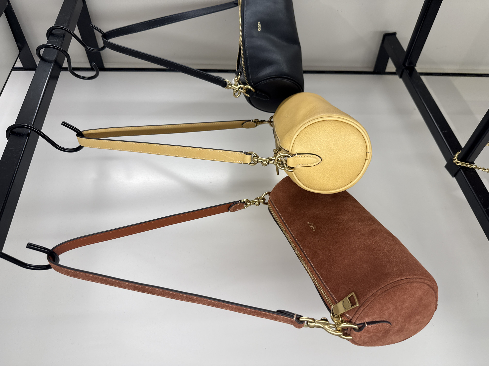 The image shows three cylindrical Coach handbags hanging from a black metal rack. The bags are arranged vertically, one above the other. The top bag is black leather, the middle bag is yellow, and the bottom bag is brown suede. The rack is located in Sunrise, United States of America.