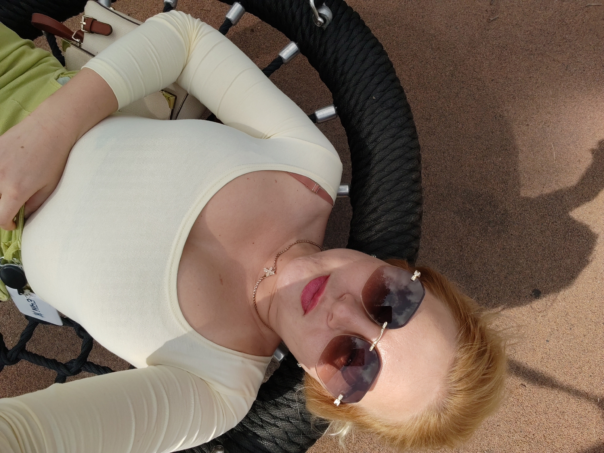 A woman lies on her back on a circular rope swing, with a brown sandy surface beneath. She wears a pale yellow, long-sleeved top, a lime green garment, and large, tinted sunglasses. Her blonde hair is visible around her face. A small purse with a brown leather strap and buckle rests near her left arm. The scene appears to be outdoors, with a shadow cast on the sand from an unseen object. The woman's head is tilted back, and her eyes are hidden by her sunglasses. Her lips are painted a bright pink. A delicate gold necklace with a small pendant is visible around her neck. The rope swing is thick and black, attached to what appears to be a metal frame. The overall impression is a relaxed, casual pose in an outdoor recreational setting, possibly a park or playground in Izhevsk, Russia.