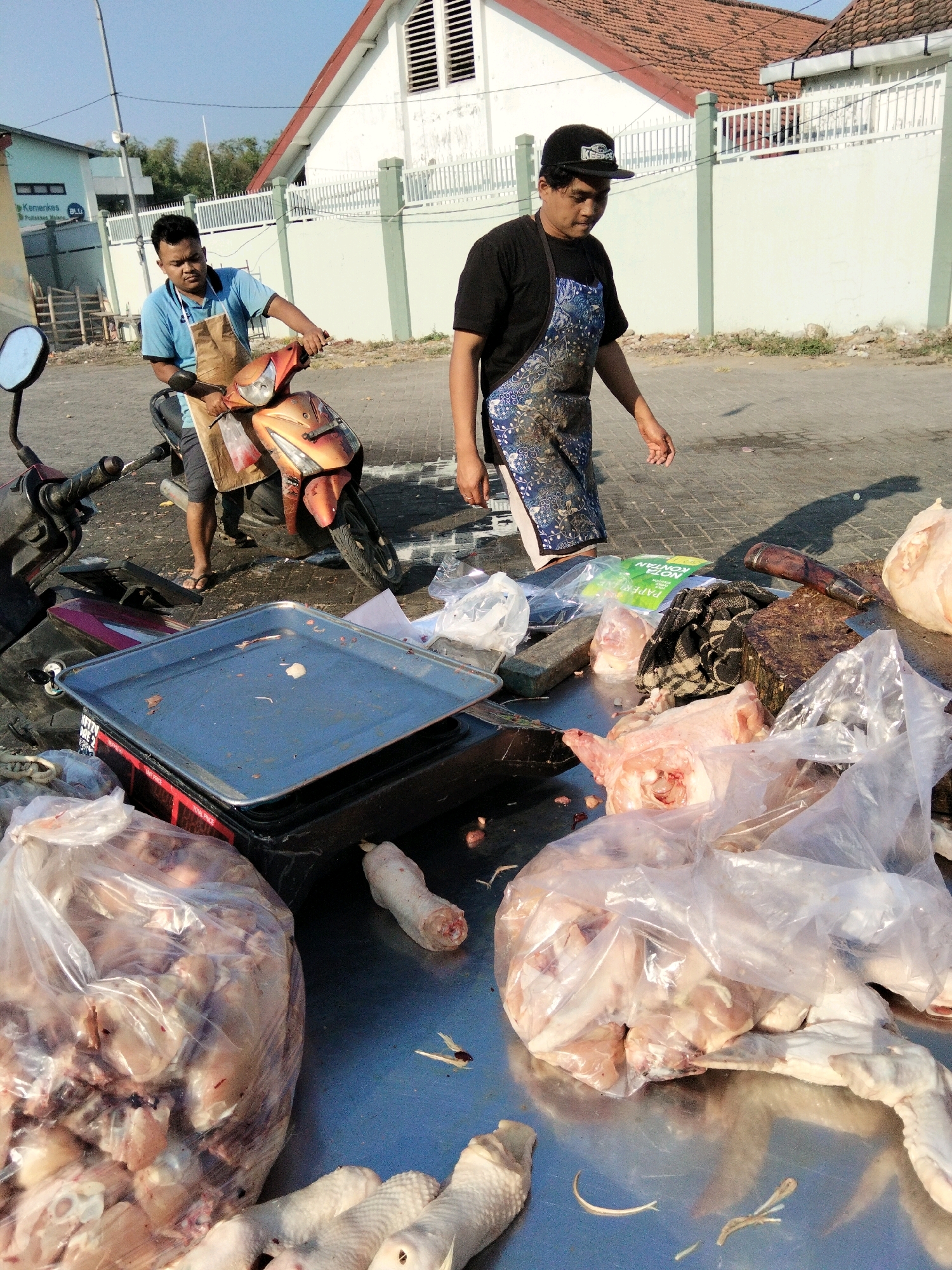 Two men are in Banyudono, Indonesia, near a building with a white facade and a terracotta roof. The man on the left, wearing a blue t-shirt and an apron, is on a motorcycle. The man on the right, wearing a black t-shirt and a patterned apron, is walking. In the foreground, there is a table with raw chicken parts, some in plastic bags and some loose. There is also a metal tray and a cleaver. The ground appears to be paved.