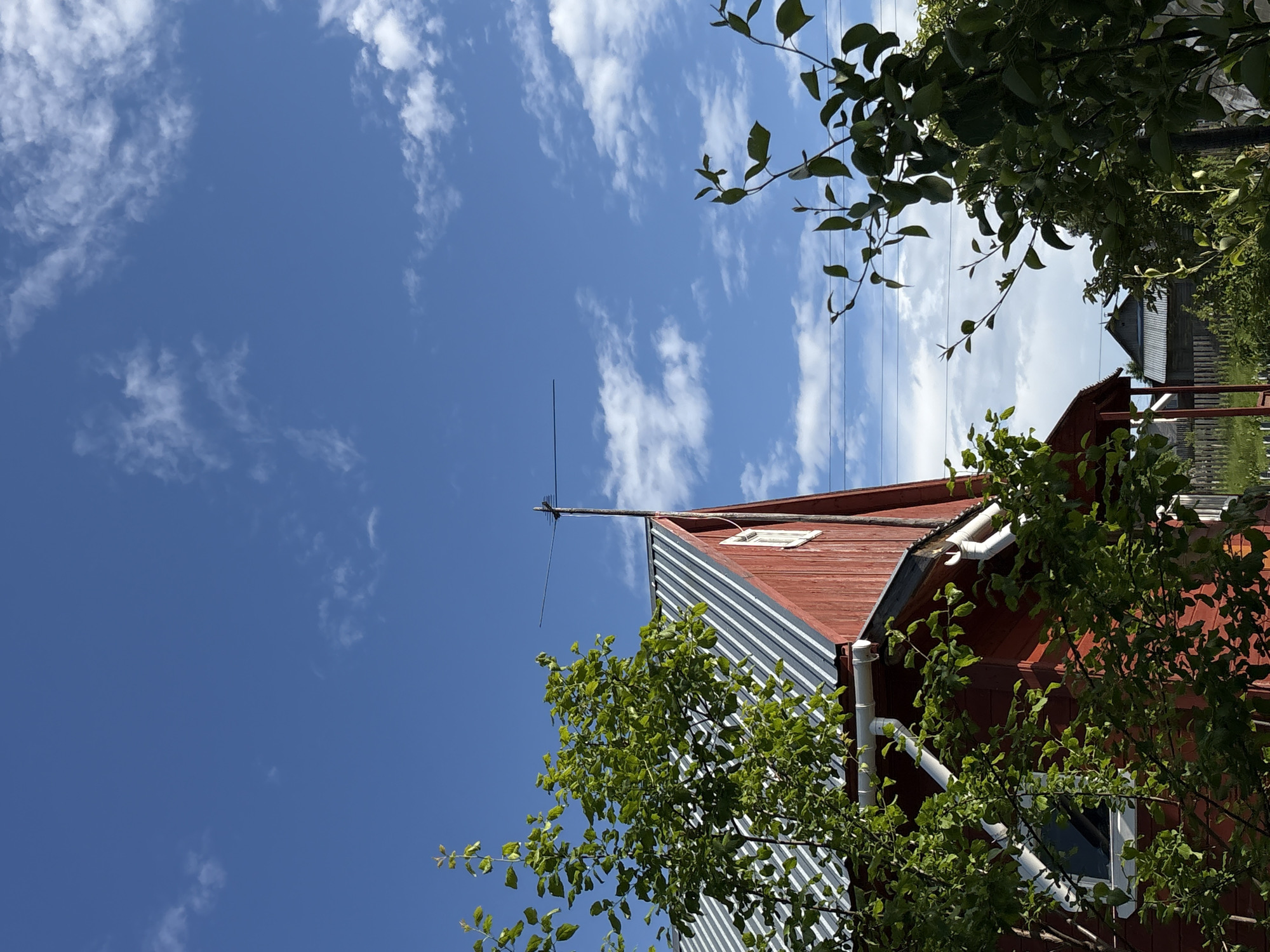 The image shows a close-up view of a red wooden house with a corrugated metal roof against a bright blue sky with scattered white clouds. A TV antenna is mounted on the peak of the roof. Green trees and foliage are in the foreground, partially obscuring the house. In the background, a wooden fence and other buildings are visible, suggesting a rural or suburban setting. The scene is set in the village of Дресвяново, Russia, on a sunny day.