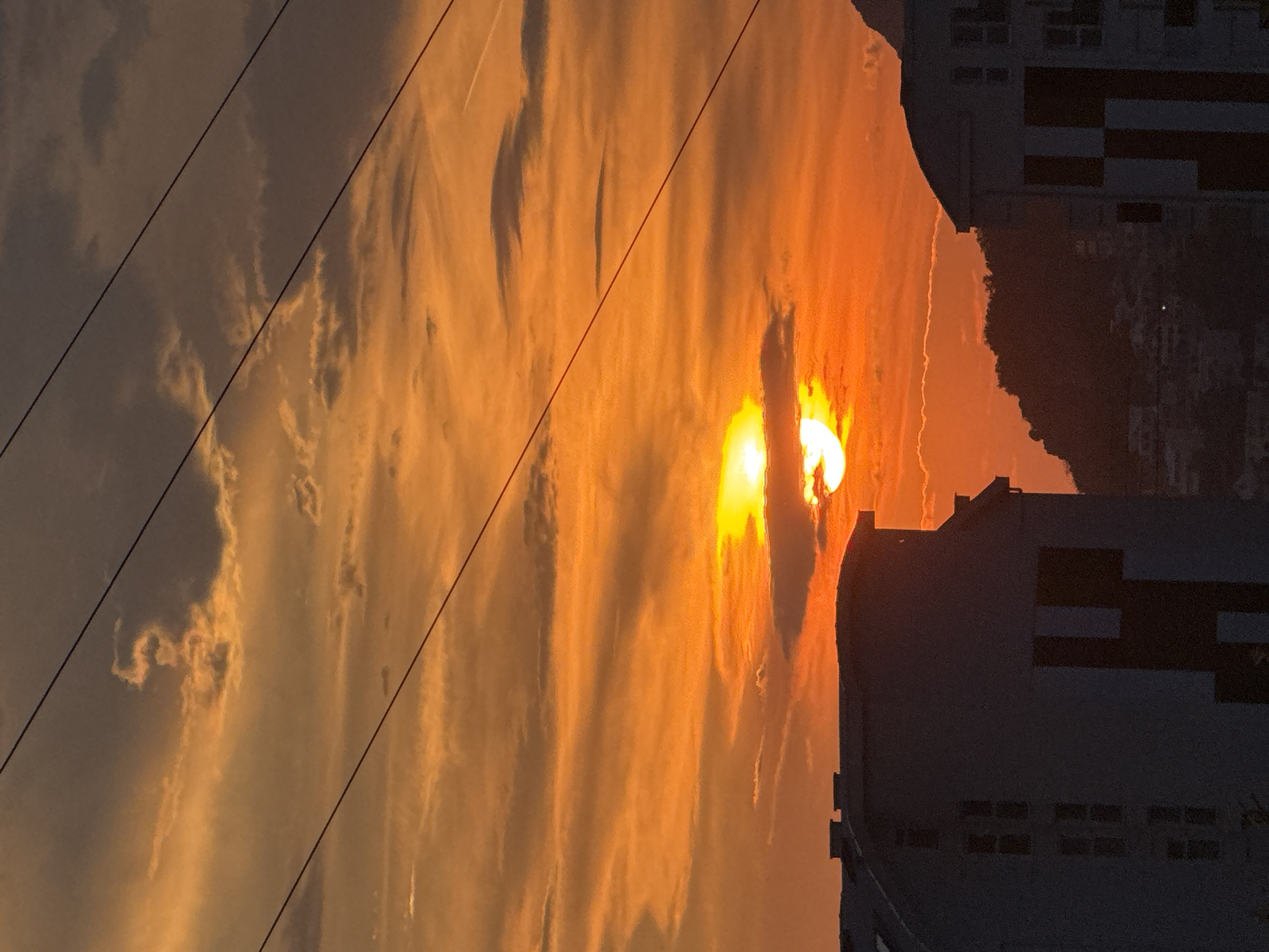 A sunset over Bar, Montenegro, features a vibrant orange sky with scattered clouds. The sun, partially obscured by clouds, is a bright orb radiating light. In the foreground, the dark silhouettes of buildings stand against the colorful sky. One building on the left has a curved facade and multiple windows. Another building on the right is also silhouetted, revealing a geometric pattern on its surface. Power lines stretch diagonally across the frame, adding a linear element to the scene. In the background, a tree-covered hill rises, with more buildings faintly visible on its slopes. The overall scene captures the dramatic beauty of a coastal sunset.