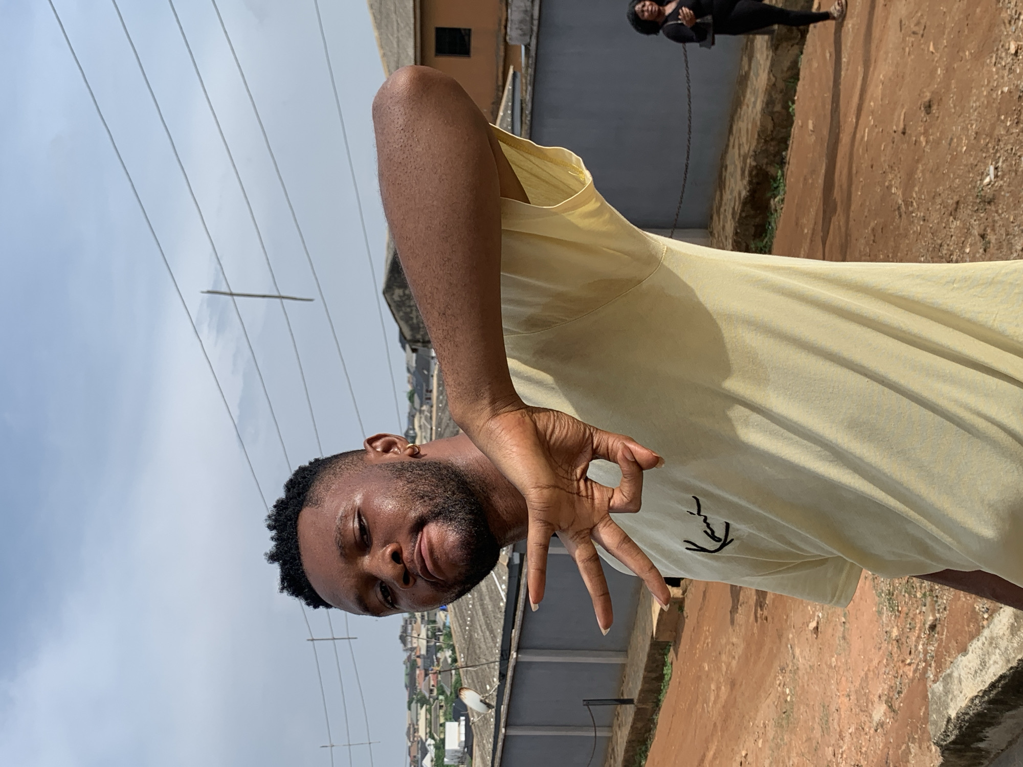 The image shows a dark-skinned man with short curly hair and a beard standing outdoors. He is wearing a light yellow t-shirt and making a hand gesture. In the background, there is a cityscape, likely in Ikorodu, Nigeria. Power lines are visible above, and another person is slightly blurred in the background standing on a raised platform with an orange-brown dirt ground. The overall environment appears to be a rooftop.