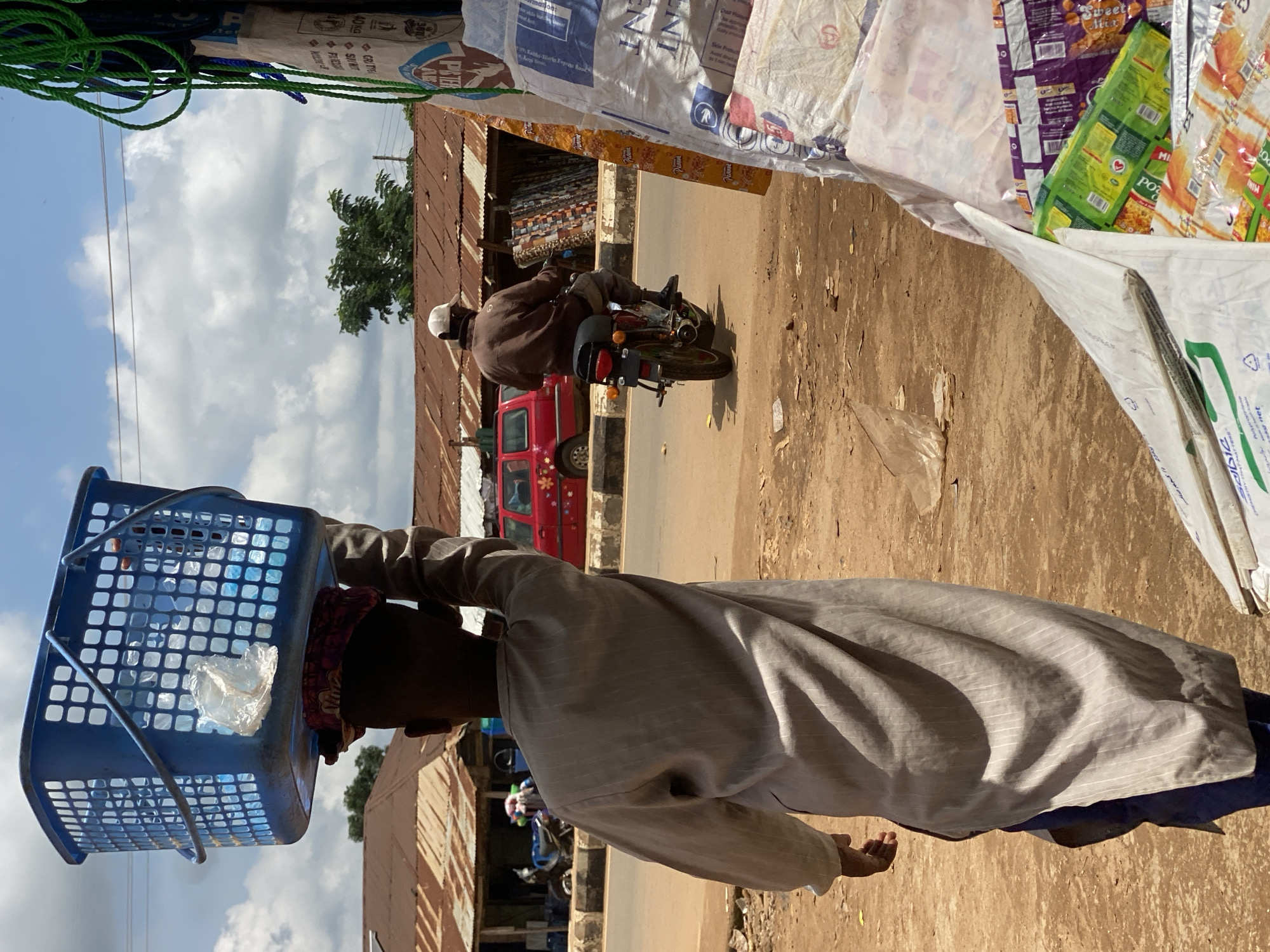 In Kafanchan B, Nigeria, a person walks away from the camera, balancing a large blue plastic basket filled with plastic water bottles on their head. They are wearing a long-sleeved, light-colored garment with vertical pinstripes and blue trousers. In the background, a man on a motorcycle rides away from the viewer on a paved road. To the right of the road are market stalls with corrugated iron roofs, displaying various goods. A red vehicle is parked near the stalls. The environment appears to be a dusty, outdoor market scene with scattered debris on the ground.