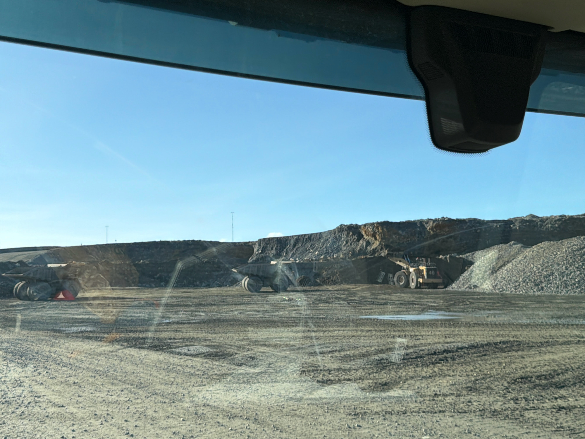 The image depicts an open-pit mine under a clear blue sky. In the foreground, a dirt and gravel surface is visible, possibly a roadway within the mine. In the middle ground, several large mining vehicles are present. On the left, a massive haul truck with multiple wheels is partially obscured by shadow and appears to be stationary. To its right, another large mining truck, a yellow wheel loader, is actively engaged, with its front bucket raised. Behind these vehicles, large piles of crushed rock or ore are stacked high, forming the operational landscape of the mine. The background consists of rugged, excavated terrain and a distant horizon under the expansive sky. The overall scene suggests a busy industrial mining operation.