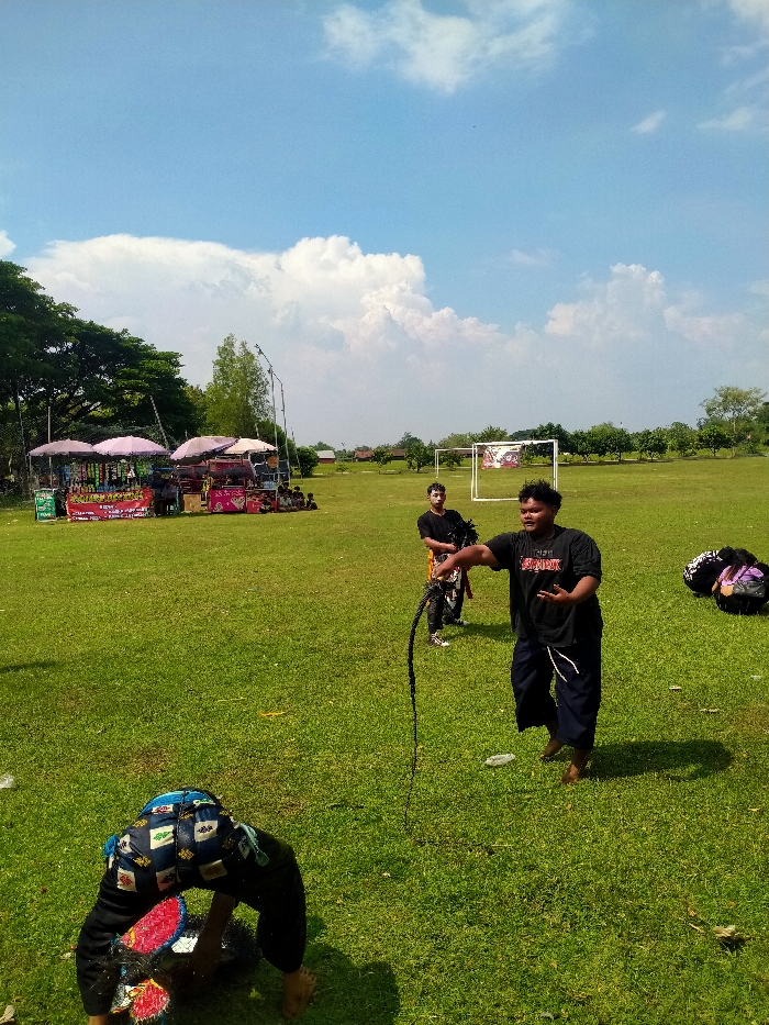 This image depicts an outdoor community event taking place on a large, sunlit green field in Jetak, Indonesia, under a clear blue sky with scattered white clouds.
In the immediate foreground, a person wearing dark pants with colorful geometric patterns and what appears to be a decorative headpiece or mask (red and black) is bent over, facing away from the camera, with their head lowered towards the grass. This individual is barefoot.
In the mid-ground, slightly to the right, a heavier-set man in a black t-shirt (with "SPIRIT" partially visible) and dark loose-fitting pants stands barefoot, holding a dark, braided whip or rope in his right hand. His left hand is open in a gesturing position, and he is looking towards the left. Behind him and slightly to his left, another man in a black t-shirt and dark pants with a yellow/gold sash or fabric around his waist stands, wearing white sneakers. He appears to be holding a dark, bundled object. To the right of the heavier-set man, a purple bag or bundle rests on the grass.
In the background on the left, a row of mature trees borders the field. Below these trees, four market stalls with light-colored umbrellas are set up, displaying various packaged goods and drinks. Several people are gathered around these stalls.
Towards the right background, a white soccer goal with a net stands on the field, featuring a colorful banner or fabric draped across its crossbar. Further back, more trees and distant low-rise buildings are visible. The grass shows some scattered litter, including plastic bottles. The scene is brightly illuminated, casting clear shadows.