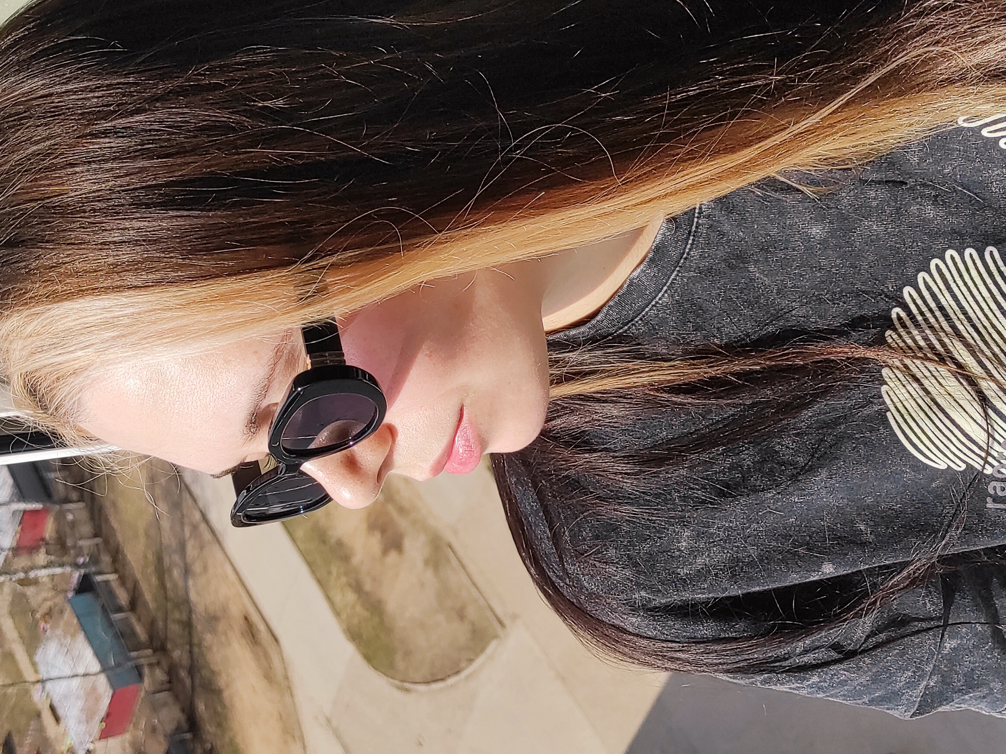 The image captures a close-up, profile view of an individual, likely female, situated outdoors under bright daylight in Chaikovsky, Russia. The person's head is tilted to the right, showing her right side from the chest up.
She has striking two-toned hair: dark brown or black at the roots and upper layers, blending into a prominent light blonde or heavily streaked blonde underneath and towards the front. She is wearing stylish black, rounded sunglasses that obscure her eyes. Her skin appears fair, and her lips are painted with a subtle light pink or nude lipstick. She is dressed in a dark grey or black t-shirt or sweatshirt made of a textured fabric. A large, light-colored graphic design is visible on the right side of her chest, depicting a radiating or fan-like pattern. The letters "ra" are partially discernible as part of a word on this graphic.
The background, somewhat blurred due to shallow depth of field, indicates an outdoor setting. Various low-rise buildings or structures, some with distinct colored roofs (such as red and blue), and areas of earthy ground are visible, suggesting a residential or semi-urban environment. The bright natural sunlight casts clear highlights on her hair and face, indicating a sunny day with clear weather. No specific activity beyond posing for a photograph or standing outdoors is evident.