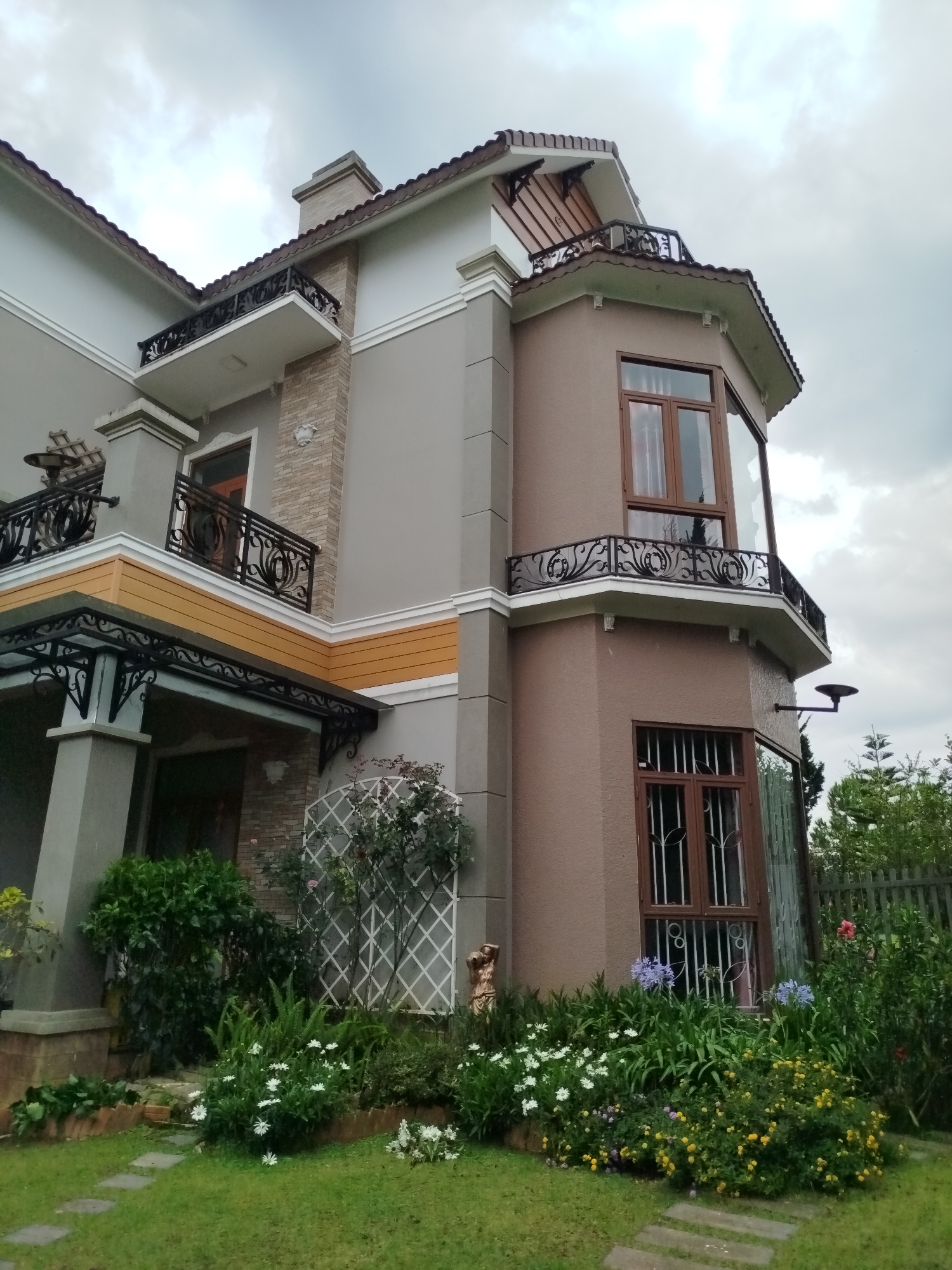 The image depicts a multi-story house in Da Lat, Vietnam. The building features a complex architectural design with different sections, including balconies with ornate black railings. The exterior walls are painted in varying shades of beige and grey, with some sections showcasing stone accents. Windows are framed in dark brown, and the roof is tiled. The house is surrounded by a well-maintained garden with a variety of plants, flowers, and a stone pathway. The sky is overcast. A small golden statue is visible amongst the plants.