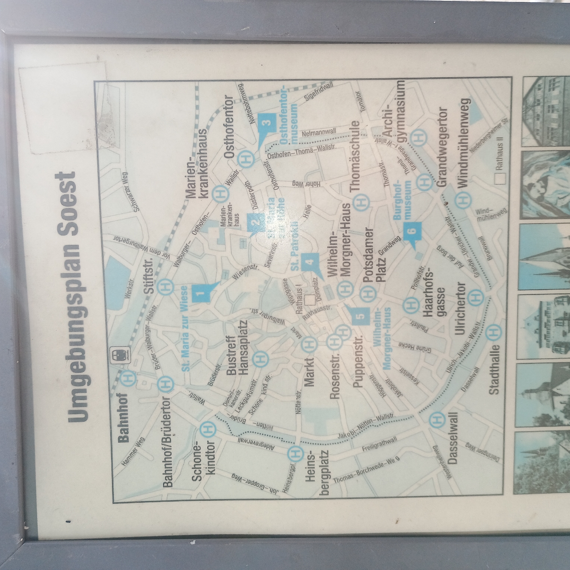 The image is a map of the surrounding area of Soest, Germany, titled "Umgebungsplan Soest". It displays a detailed city map with street names, landmarks, and points of interest marked with blue circles containing the letter 'H', likely indicating parking or transport hubs. The map is enclosed in a gray frame and appears to be mounted outdoors, possibly near a public transportation stop or tourist information point. Several numbered blue squares on the map indicate specific locations: "1. St. Maria zur Wiese", "2. St. Maria zur Höhe", "3. Osthofentor-museum", "4. St. Patrokl", "5. Wilhelm Morgner-Haus", and "6. Burghofmuseum". Below the main map, there are smaller inset images depicting architectural structures, likely historical buildings in Soest. The overall scene is static and informative, serving as a guide for navigating the city and its attractions.