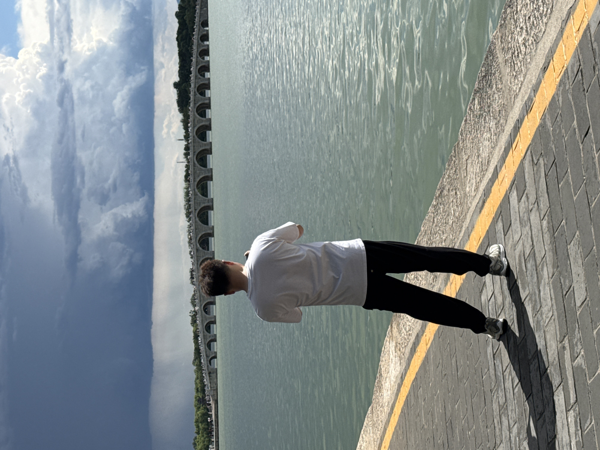 A lone individual, viewed from the back, stands on a dark gray paved walkway with a prominent yellow stripe delineating its edge. The person is a male, wearing a white short-sleeved t-shirt, black trousers, and light-colored athletic shoes. He is holding a small electronic device, presumably a phone or camera, with both hands, pointed towards the distant scene. In the mid-ground, a vast expanse of calm, greenish-blue water, identified as Kunming Lake, extends to a multi-arched stone bridge, specifically the Seventeen-Arch Bridge, located within the Summer Palace in Haidian District, Beijing, China. Small figures of people are visible on the bridge. Beyond the bridge, a tree-lined landmass is apparent. The sky displays contrasting weather phenomena: dark, heavy rain clouds with visible rain streaks are present on the left side, while lighter, fluffier clouds and patches of blue sky are observed on the right.