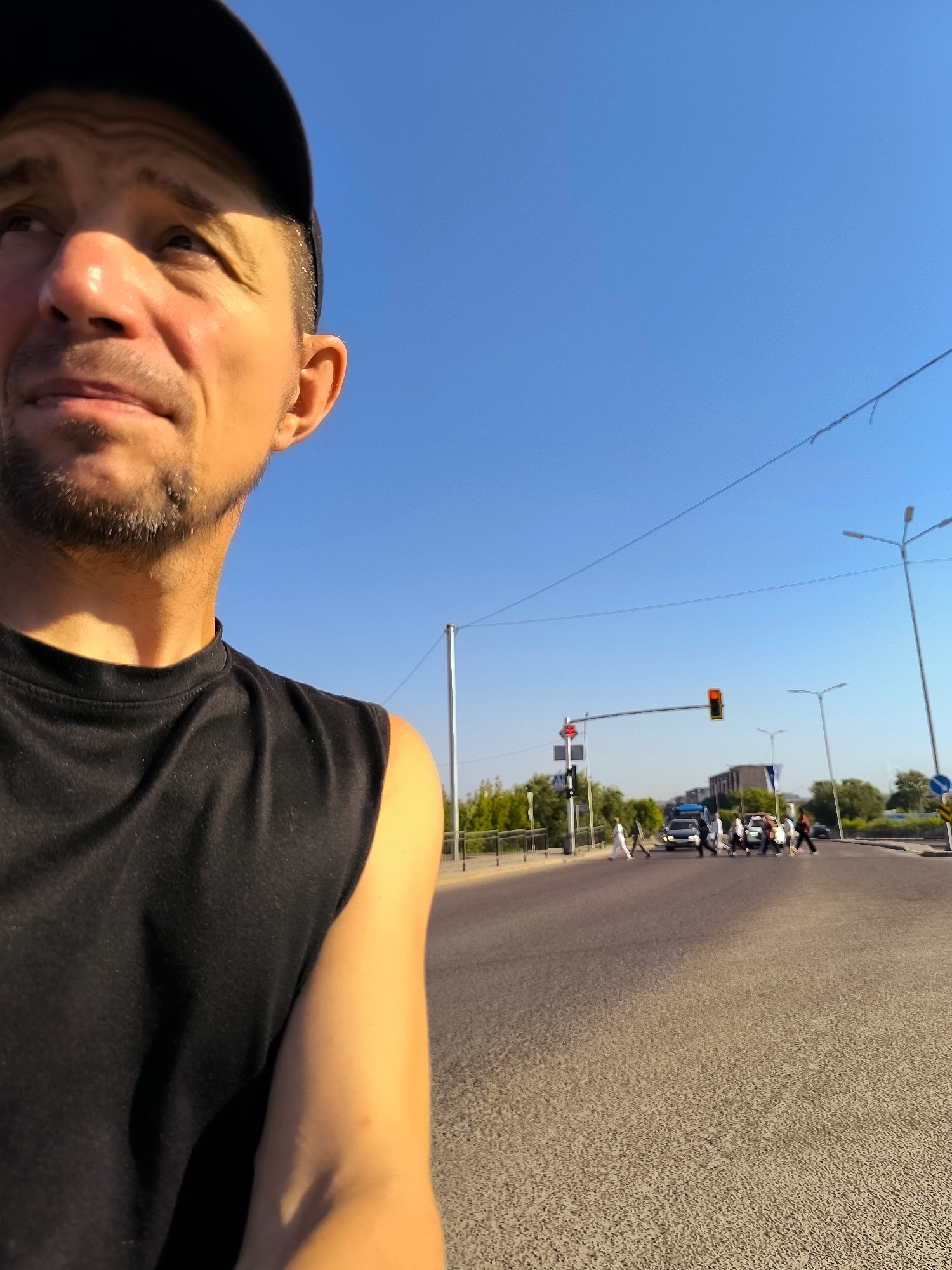 In an outdoor urban scene under a clear blue sky, a man wearing a black baseball cap and a black sleeveless shirt is captured from the chest up on the left side of the frame. His face, partially obscured by the cap, is turned upwards and to the left, with sunlight highlighting his features and the side of his face. In the background, a street intersection is visible. A traffic light shows a red signal. Pedestrians are crossing the street in a crosswalk, and several vehicles are stopped at the intersection. Trees line the side of the road, and some buildings can be seen in the distance. The setting appears to be a city, likely Qarağandı city administration, Kazakhstan, given the context provided.