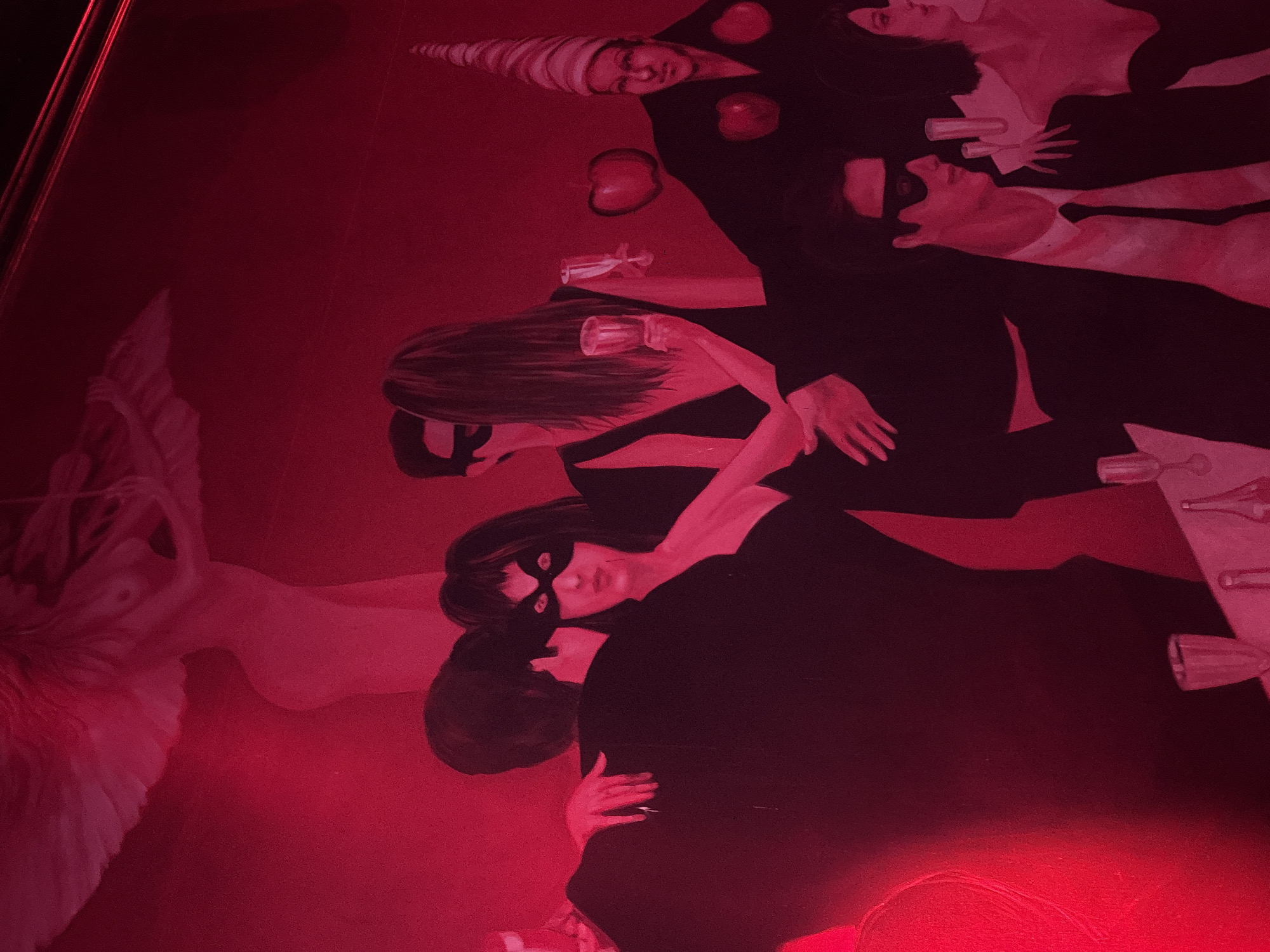 A mural or large painting depicts a masked party scene in Mexico City, Mexico, illuminated by pervasive red lighting. In the upper left background, an ethereal, winged female figure with outstretched arms and a flowing form is rendered against the deep red wall. The midground features several individuals. To the left, a man from behind embraces a woman wearing a black eye mask, who looks directly forward. Adjacent to them, another couple is depicted; a man with his back to the viewer stands close to a woman in a black dress who holds a stemmed glass and faces away. To the right, a figure wears a black robe or suit, a conical striped hat, and has two prominent red apples affixed to their chest and shoulder. This figure also appears to be holding an arm out, interacting with the central group. Above this figure, a solitary red apple floats against the wall. In the lower right foreground, a man in a black suit, white shirt, and black tie wears a black eye mask and is seated at a table. His right hand is visible on the table, where several wine glasses and bottles are present. A partially visible woman in a black dress is seated to his right. The entire scene is bathed in a strong, reddish-pink hue, suggesting a theatrical or intimate atmosphere.