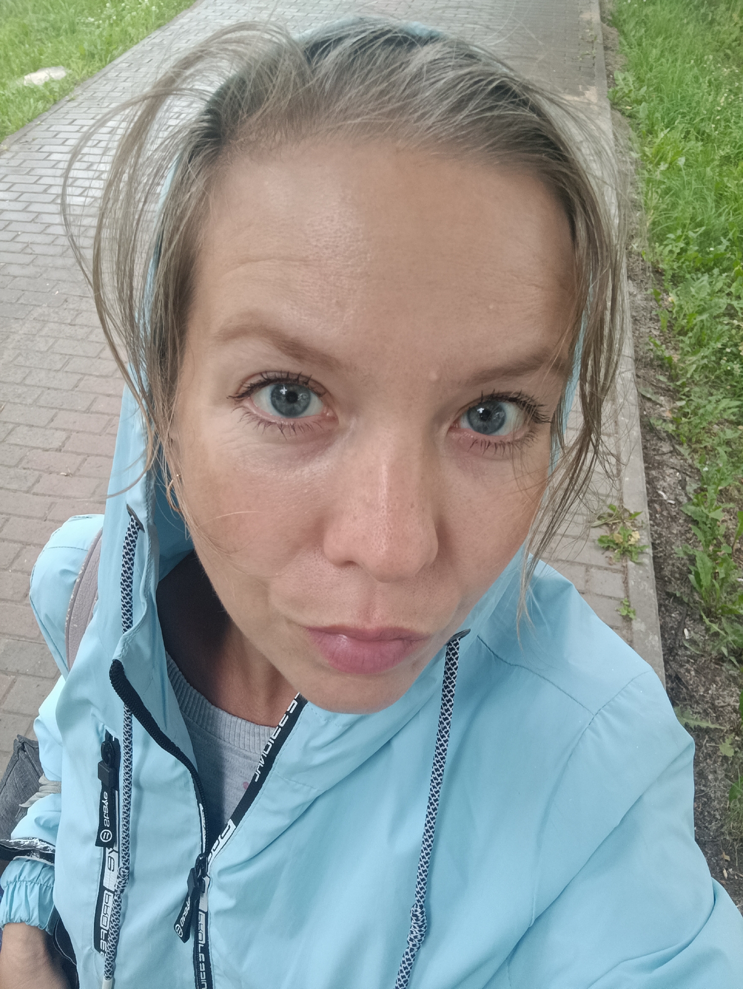 A selfie taken by a Caucasian woman with blonde hair and blue eyes. She is wearing a light blue hooded jacket over a grey sweater. The woman is looking at the camera with a neutral expression. The background appears to be an outdoor setting, possibly a park or a sidewalk, with a paved path and green grass. There are no other people or objects of interest visible in the image. The location is identified as Chaikovsky, Russia.