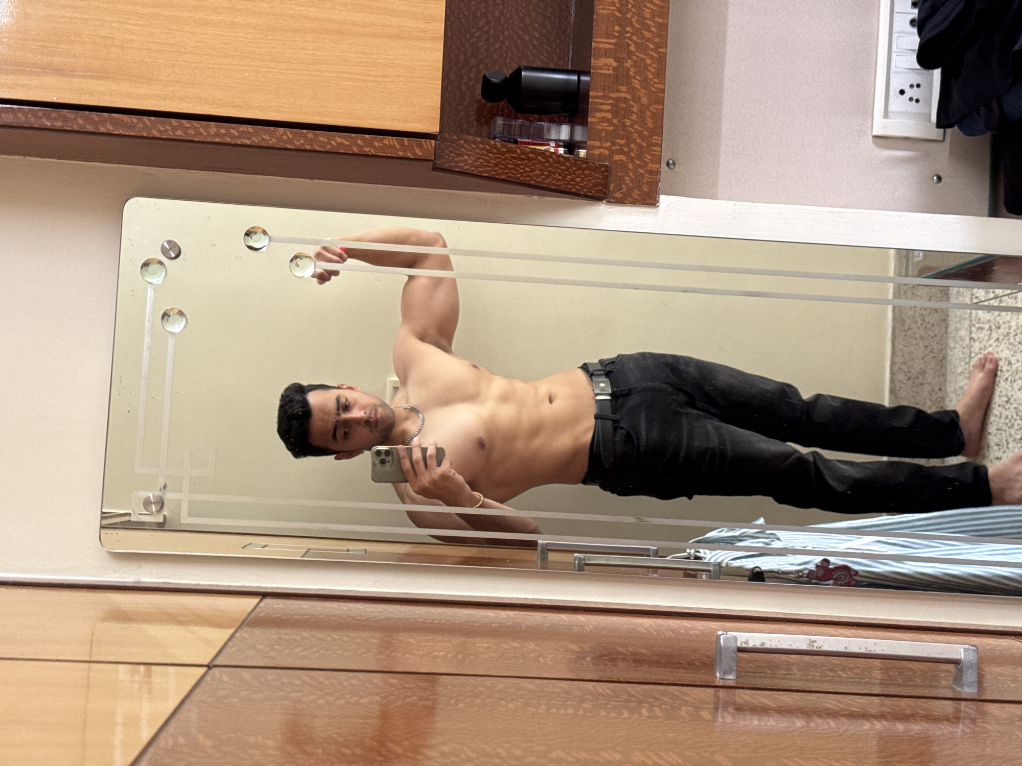 A male reflects in a mirror, taking a selfie with a smartphone. He is bare-chested, wearing black pants and accessories. The mirror is framed and reflects a room with an outlet, clothes drying on a rack, and wooden furniture. The location appears to be Kolkata, India.