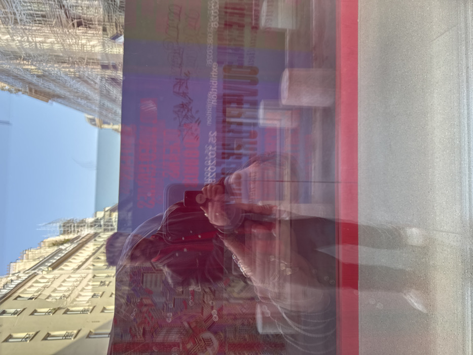 The image presents a layered scene composed of a reflection on glass superimposed over a Parisian streetscape.
A male individual is captured in a blurred reflection, visible primarily from the waist up. He wears a dark jacket over a red inner garment and holds a black smartphone with a white pop socket in both hands, positioned as if taking a photograph or video. Rings are visible on his right hand. His lower body is further blurred, showing white shoes. A prominent horizontal red stripe traverses the lower third of the reflection.
Through the glass, a street in Paris, France, is visible under a clear blue sky. On the left, a typical multi-story Haussmannian building with light-colored facades and numerous windows lines the street. On the right, another similar building is heavily obscured by extensive white scaffolding, indicating construction or renovation work. A large, blue-purple banner is displayed horizontally across the mid-ground, featuring mirrored and distorted French text, including "exposition" and dates "22.10.2021-22.01.2022," suggesting an advertisement for an exhibition. Several light-colored cylindrical bollards or barriers are visible on the ground, behind the banner's lower edge. The architectural style and French language confirm the location as Paris.