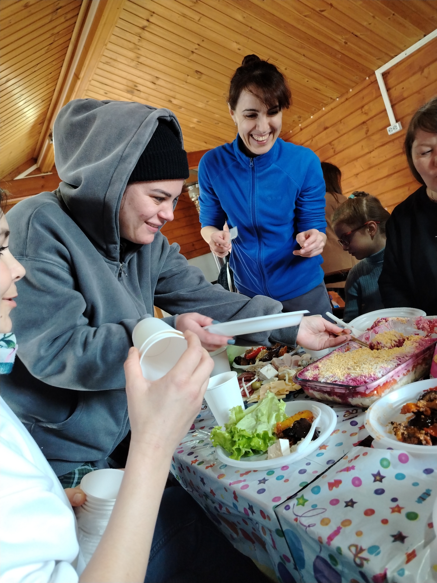 The image shows a group of people gathered around a table laden with food, likely indoors given the wooden ceiling. A woman in a grey hoodie and black beanie is serving food from a dish onto a white plastic plate. Another woman in a blue tracksuit is standing and smiling, appearing to interact with the group. The table is covered with a patterned tablecloth featuring stars and polka dots. The food includes a layered salad in a glass container, as well as other dishes and salads in white plastic plates and bowls. White plastic cups are also visible. A girl with glasses can be seen in the background. The location and city are undetermined based on the image.