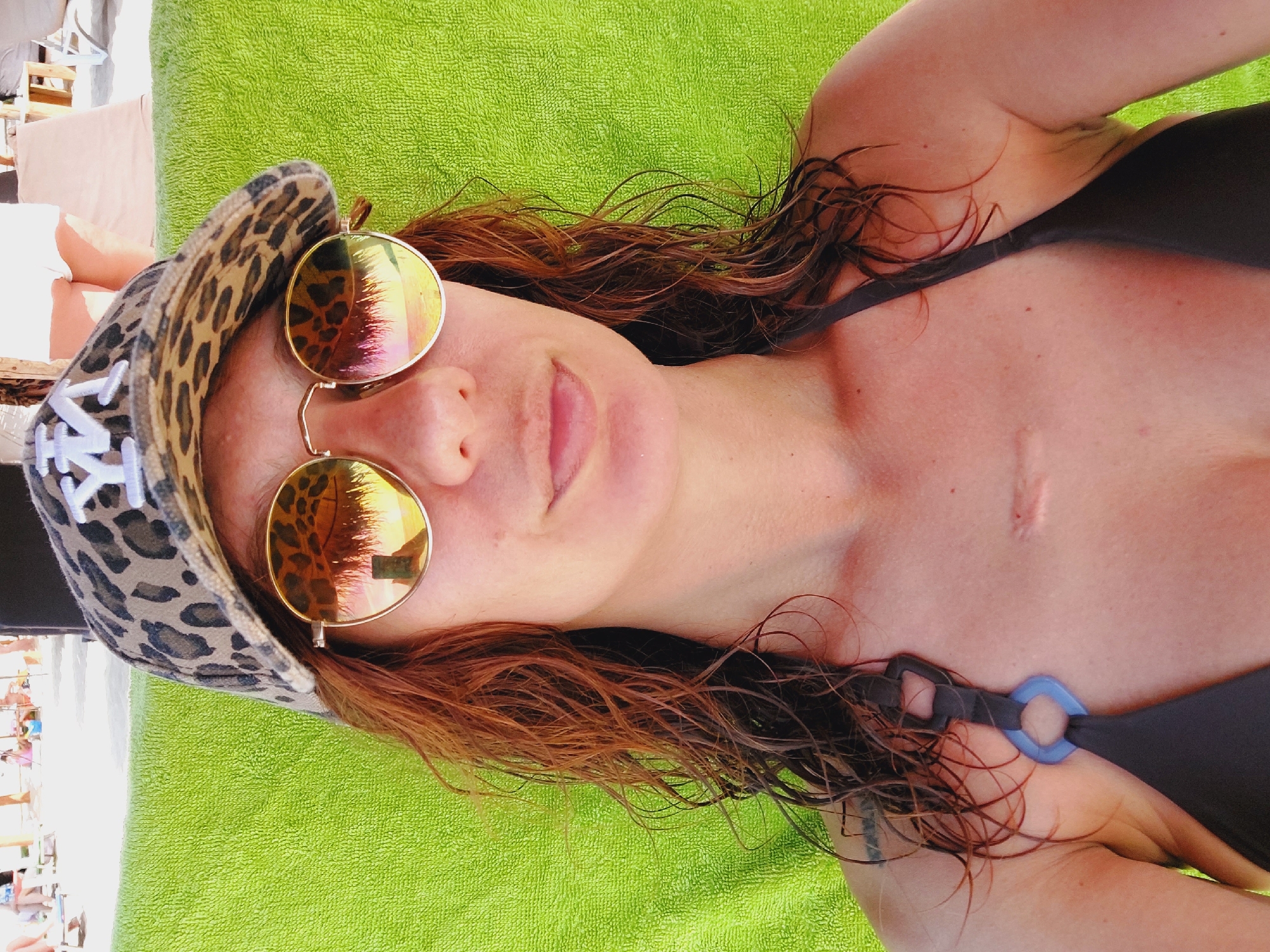 The image is a close-up selfie of a woman wearing a leopard-print baseball cap with a white logo, round gold-rimmed sunglasses with reflective lenses showing a beach scene, and a dark bikini top. Her hair is wet and reddish-brown, falling loosely around her face and shoulders. She is resting on a bright green textured surface, likely a beach towel or chair. A scar is visible on her upper chest. The background shows a blurred beach environment with people and beach furniture. The scene is set in Sozopol, Bulgaria.