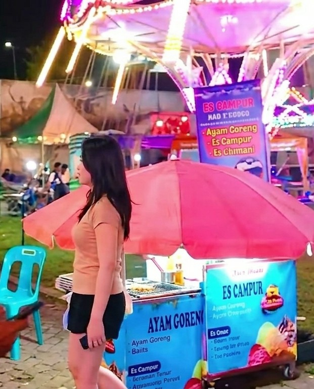 The image depicts a scene in Medan, Indonesia, likely at a night market or fair. A young woman with dark hair is walking past two food stalls, one selling Ayam Goreng (fried chicken) and the other Es Campur (a mixed ice dessert). Both stalls are under pink umbrellas and have blue signage. The background features other food vendors and carnival rides, illuminated by colorful lights. People are seen in the distance, suggesting a bustling environment.