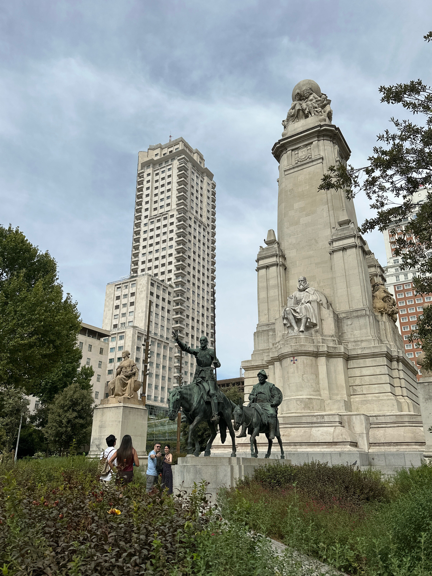 The image shows the Plaza de España in Madrid, Spain. In the foreground, there is dense green foliage with some reddish-brown bushes. Behind the foliage, there are several people. One person, facing away from the camera, has dark hair and is wearing a dark red top and dark pants. Another person, also facing away, is wearing a white top and light-colored shorts, carrying a backpack. To the right, a couple is taking a selfie. The man is wearing a blue t-shirt and grey shorts, and the woman is wearing a black and white polka dot dress.
Dominating the middle ground and background are several prominent structures. To the right is the Monument to Cervantes, a large, stone monument featuring sculptures. A notable sculpture on the monument depicts a man seated with a book, and at the very top, a sphere is held by figures. In front of the monument are two bronze equestrian statues. The one on the left appears to be a knight on horseback, possibly pointing. The one on the right depicts another figure on horseback.
Behind the monument and statues, two tall, modern buildings are visible. The taller one, on the left, is a light-colored skyscraper with many floors and balconies. To its right, and slightly behind, is another, slightly shorter light-colored building. Further in the background, parts of other buildings are visible, including one with reddish-brown brickwork. The sky above is overcast with a light grey, cloudy texture. Trees with green foliage are present on the left side of the image, framing the scene.