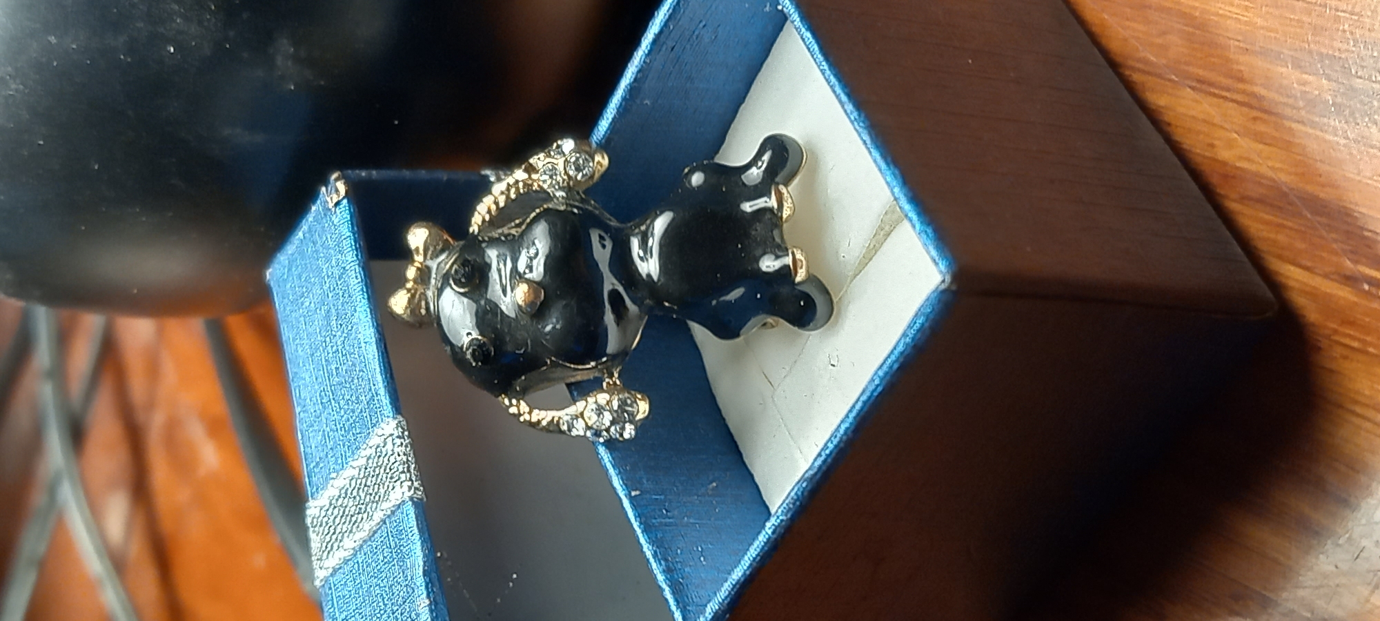 A decorative black and gold-toned object, possibly a brooch or small figurine, lies partly within a small blue textured box with a silver-colored ribbon. The object is shaped like a stylized fish or animal, featuring a glossy black body accented with gold-colored elements and small clear, sparkling stones, particularly on its fins and tail. Small gold-colored details and black dots are also visible on its body.
The blue box, possibly a jewelry or gift box, has a ridged or woven texture and a shiny silver-colored ribbon detail wrapped around one corner. The inside of the box is lined with a lighter, possibly white, material that shows some creasing.
The box and object are resting on a wooden surface with visible grain patterns, indicating a natural wood material. In the background, a dark, indistinct, possibly rounded object is partially visible on the left, while the wooden surface extends to the right. The lighting suggests an indoor setting, with no specific time of day or weather cues evident. There is no visible text in the image. The location context provided for the image is Kryvyi Rih, Ukraine.
