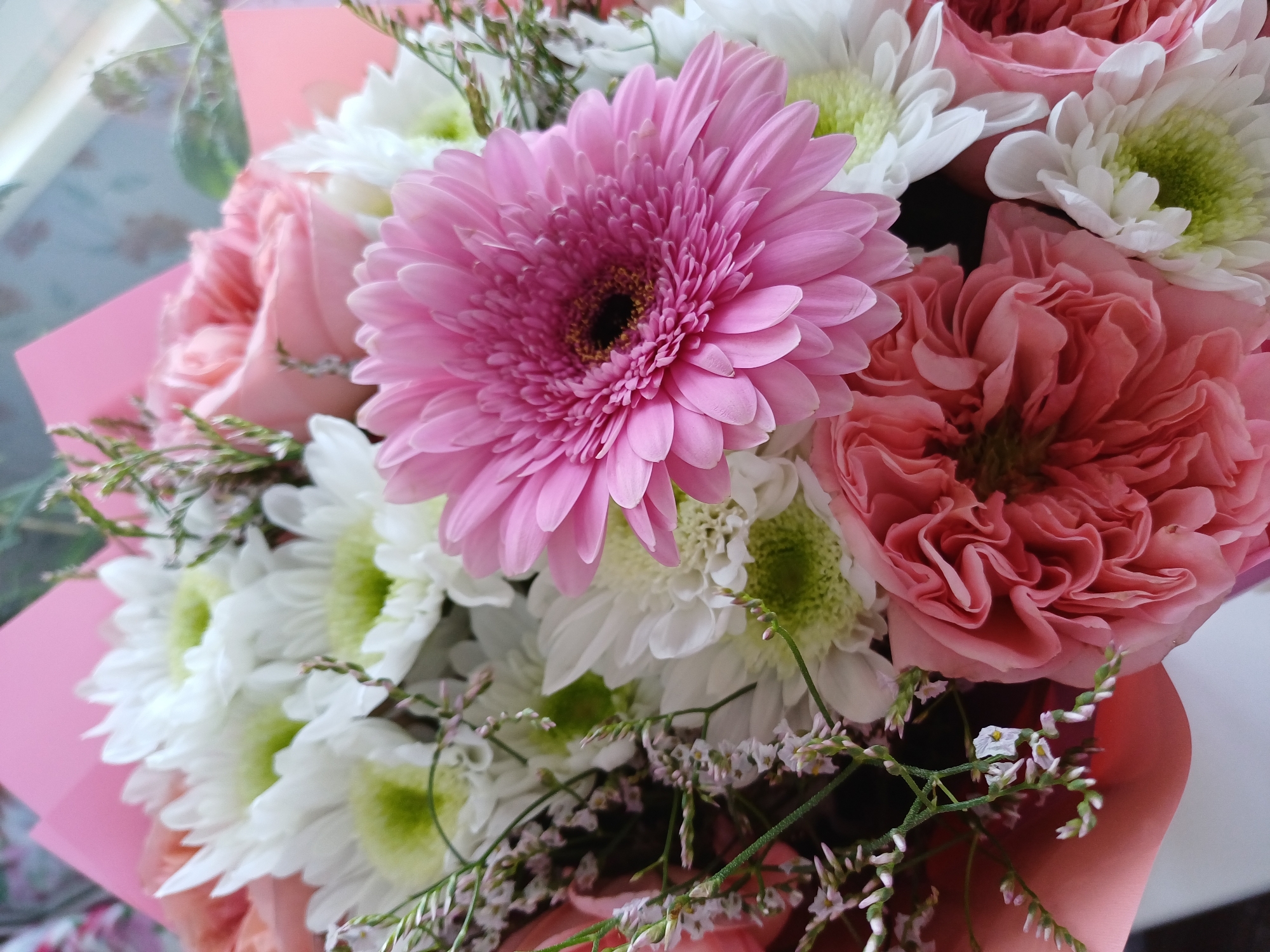 A close-up, overhead shot displays a bouquet of various flowers. The bouquet features a large pink gerbera daisy in the foreground, with its petals unfurled. Behind it, several white chrysanthemums with light green centers are visible. Also present are two large, pink, ruffled roses, one in the lower left and another peeking from the right. The flowers are arranged with some delicate, light pink filler flowers and greenery. The bouquet is partially wrapped in pink paper. The background is blurred, with hints of a patterned surface and a light source.