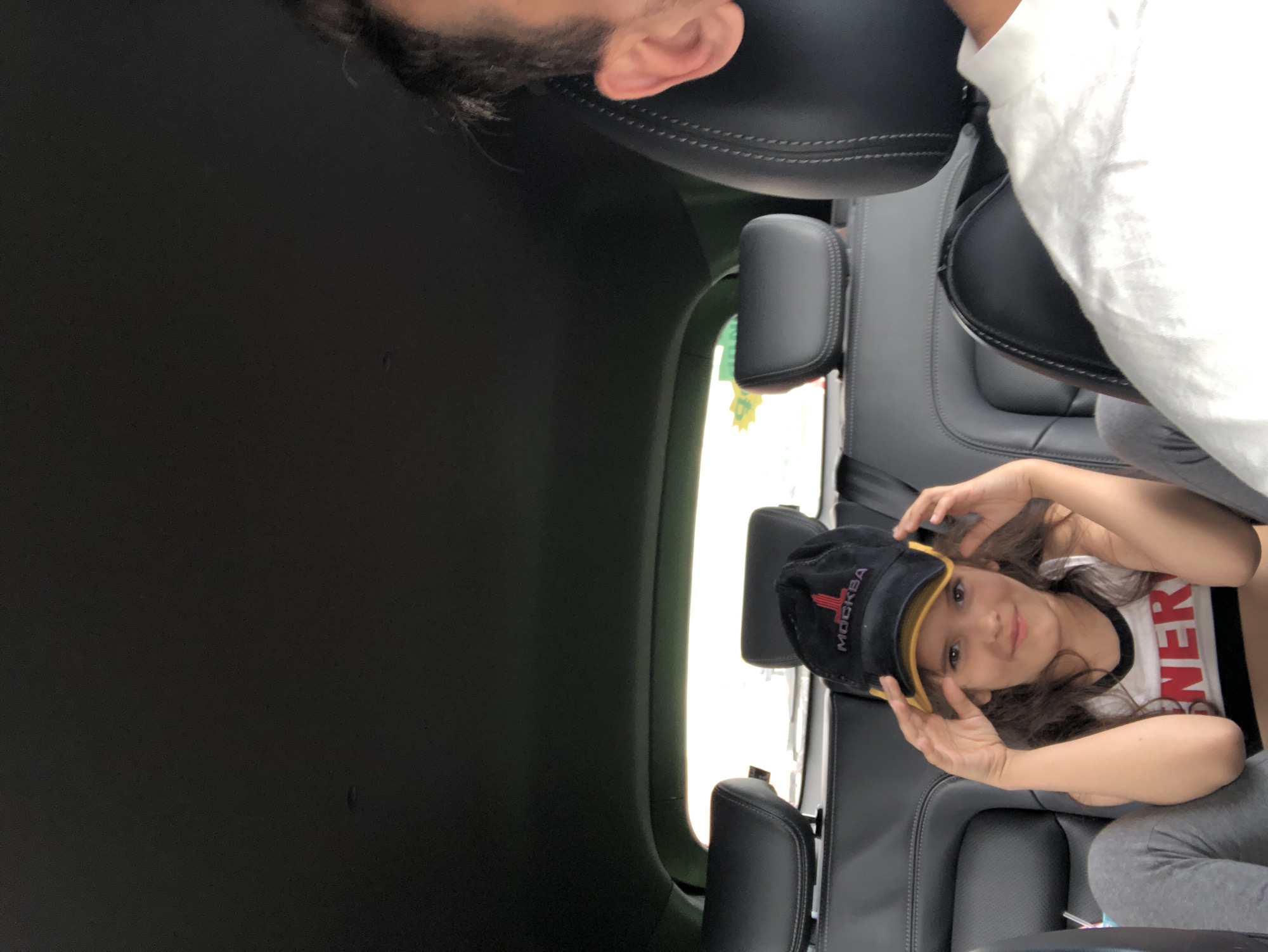 The image depicts a young girl with long, brown hair lying on her back in the back seat of a car. She is wearing a black baseball cap with a yellow brim and the word "МОСКВА" (Moscow) in red lettering on the front. She is also wearing a white t-shirt with red lettering that appears to say "SINERGY". Her hands are resting on the sides of her head, and she is looking at the camera with a slight smile. The interior of the car is visible, with dark leather seats and a window showing an outdoor scene. The lighting suggests it is daytime. The car appears to be in a city setting, potentially Saint Petersburg, Russia, given the provided location context.