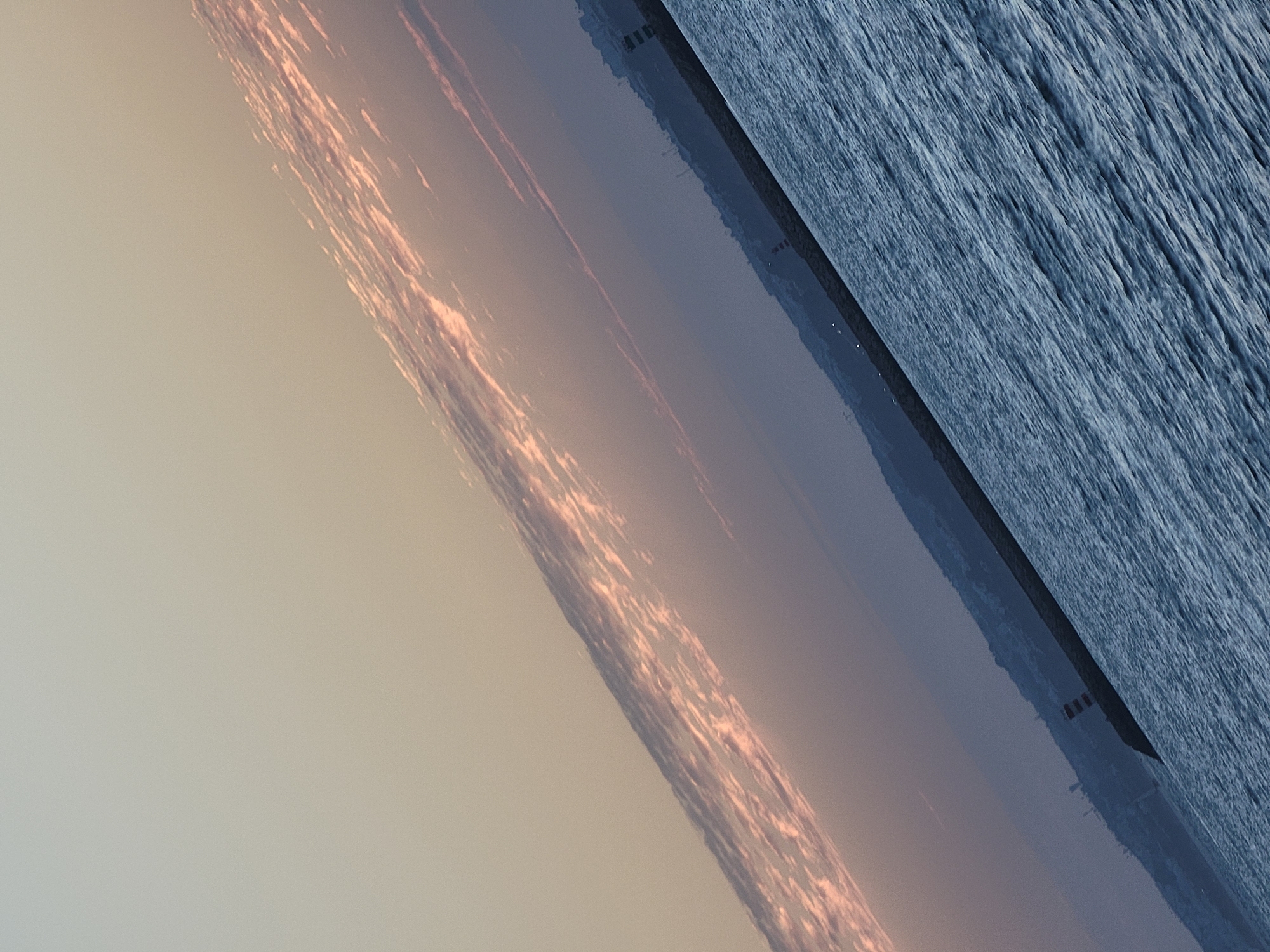 The image depicts a body of water, likely the Atlantic Ocean, under a twilight sky. The water exhibits a textured surface with visible ripples. In the distance, a coastline is visible, featuring a town or city with buildings silhouetted against the horizon. A prominent breakwater or jetty extends into the water, with a lighthouse or navigational marker present on it. The sky displays subtle color gradients, transitioning from a light, muted tone at the top to darker hues closer to the horizon. Wispy clouds, tinged with pink and orange, are present in the upper portion of the sky, suggesting either sunrise or sunset. The orientation of the image is tilted. The scene is likely located in the vicinity of Quarteira, Portugal, given the presence of a coastal town and a breakwater with a lighthouse, which are characteristic features of such locations. There are no people or discernible actions depicted in the image.