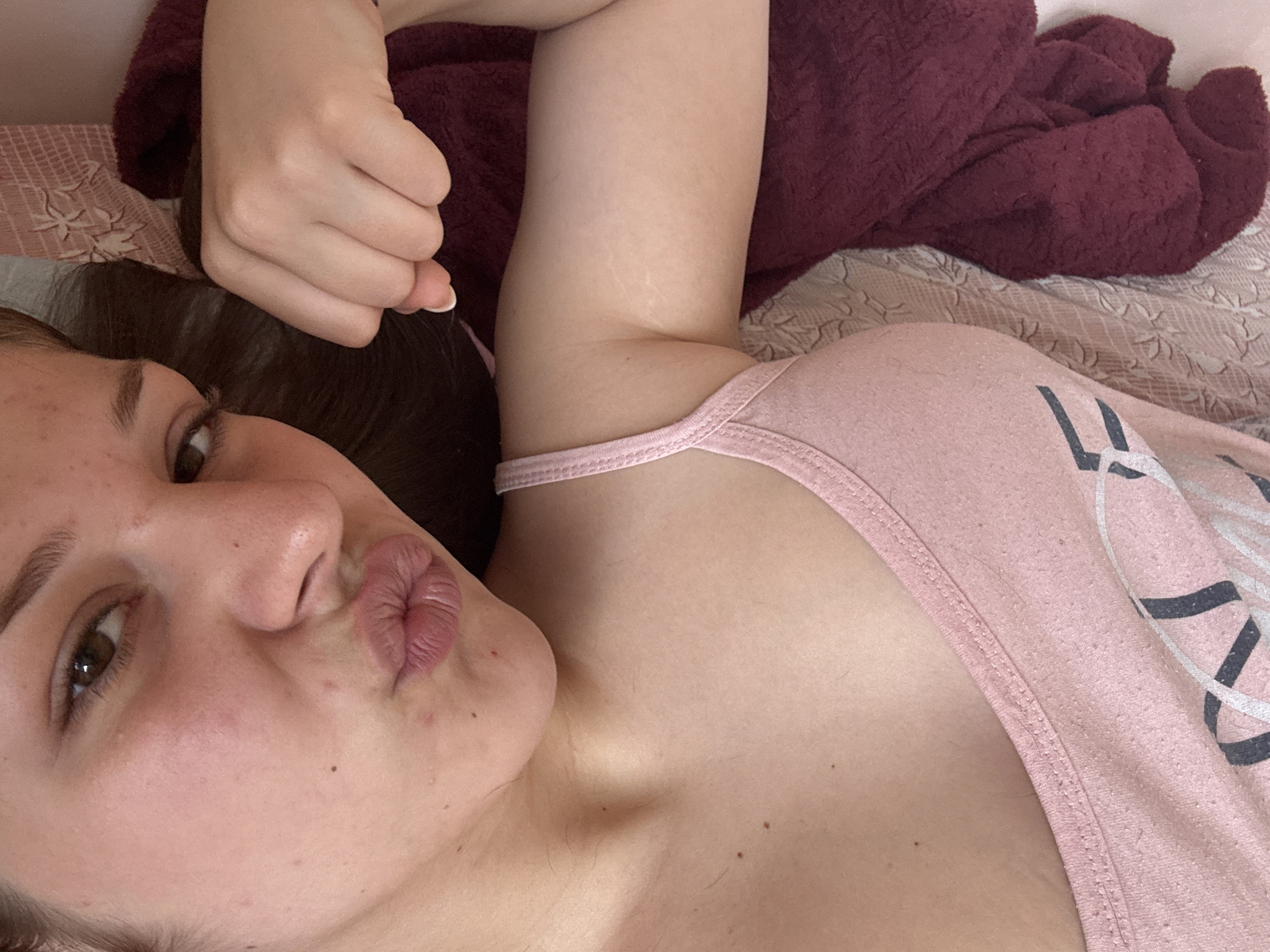 A young woman is lying on a bed, looking towards the camera with her lips pursed in a kiss. She has light skin and brown eyes. She is wearing a pink tank top. Her right arm is bent and her hand is resting near her head, which is partially covered by a dark red blanket. The bed has a light-colored patterned sheet. The lighting suggests it may be daytime, possibly indoors. There is no visible text in the image.