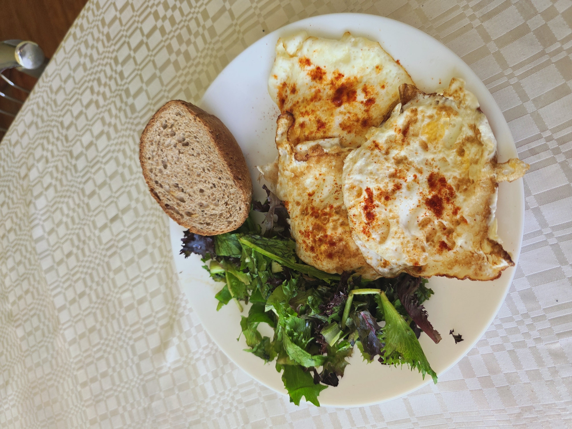 A plate of food rests on a light-colored, patterned tablecloth. The plate contains three fried eggs, dusted with red spice, a small salad with mixed greens, and a slice of whole-wheat bread. The eggs appear to be cooked sunny-side up, with visible yolks. The salad consists of various green and reddish-purple leaves. The bread is dark and has a slightly rough texture. The background shows a continuation of the patterned tablecloth, suggesting an indoor setting, possibly a dining area. There are no people visible in the image. The lighting appears natural, casting subtle shadows on the tablecloth. The scene depicts a simple meal served on a white plate.