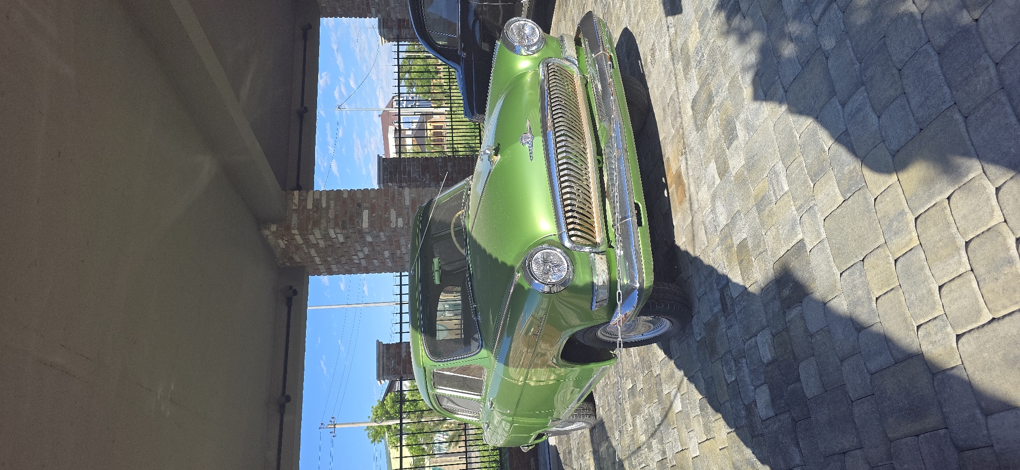 A green GAZ-21 Volga car is parked outdoors on a paved area made of interlocking gray stones. The vehicle is positioned beside a building with a concrete ceiling and brick accents, providing partial shade. In the background, a fence and glimpses of buildings under a blue sky are visible. The location appears to be Unknown, Unknown.