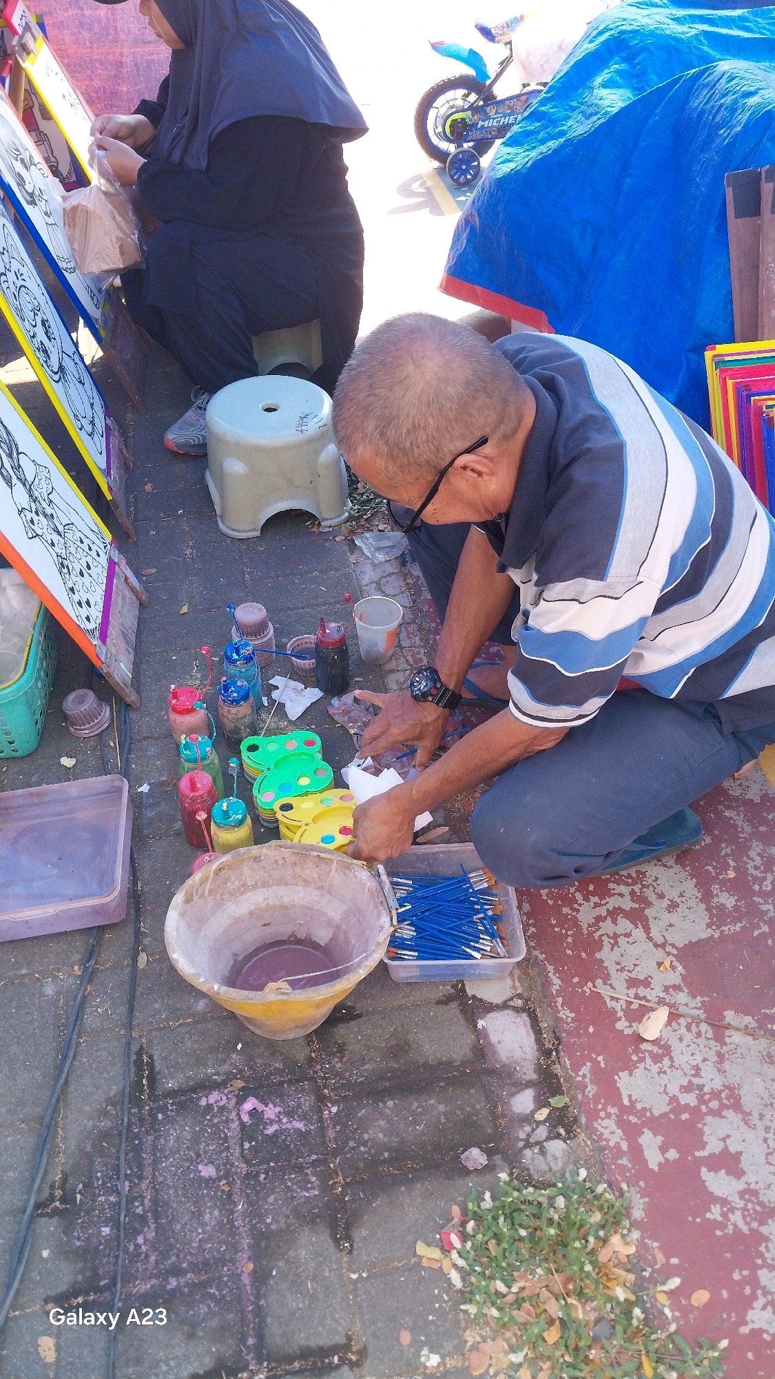 The image captures an outdoor scene, possibly a street or market stall, with a focus on art supplies and a man engaged in painting. The setting appears to be an urban environment, as suggested by the paved ground and the presence of a bicycle in the background. The man, wearing a striped shirt and dark pants, is kneeling and actively painting or preparing his materials, with a water bucket and brushes in front of him. A woman in a hijab is also visible, seated and seemingly involved in preparing art materials. Various bottles of paint, color palettes, and drawings are spread around. The presence of a "Galaxy A23" watermark indicates the photo was taken with a Samsung Galaxy A23 smartphone. The overall impression is of an artist's workspace at a public event or a street vendor.