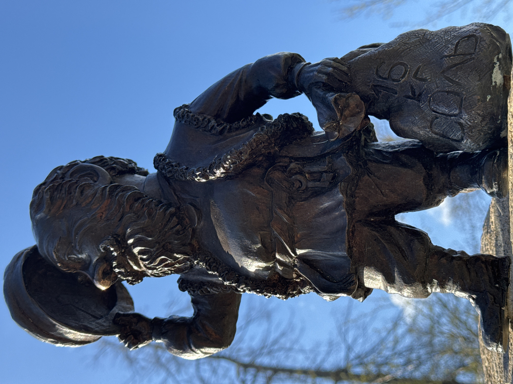 A bronze statue depicts an elderly man with a beard and curly hair, wearing a fur-trimmed coat. He carries a bowl in one hand and a sack labeled "Коляда" in the other. The statue is positioned against a backdrop of a clear blue sky and some bare tree branches, suggesting an outdoor setting. This statue is located in the city of Cherepovets, Russia.