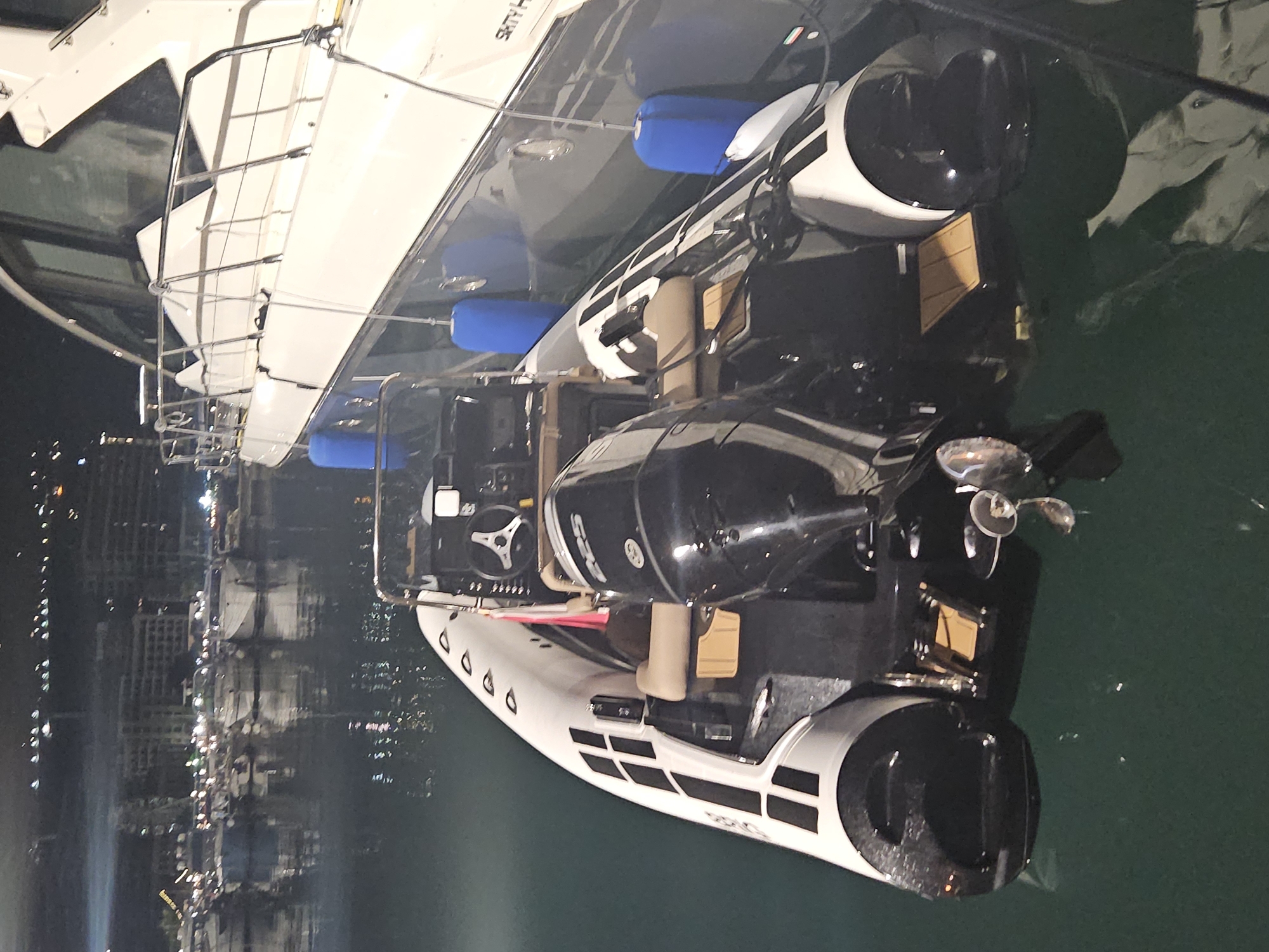 In an unknown location, a black and white inflatable boat with a large black outboard motor is docked next to a larger white yacht. The inflatable boat has tan seating and a steering console with a black steering wheel. The outboard motor has "325" visible on its cowling. Blue fenders are visible on the side of the yacht, and the yacht has a railing. In the background, a city skyline with buildings and lights is visible across the water, reflecting on the dark, calm surface. The scene is illuminated by artificial lights.