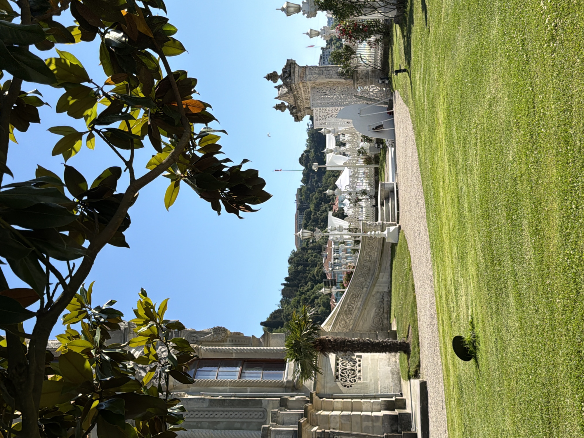 The image depicts a sunlit exterior view of a grand building with ornate architectural details, likely a palace or mansion, situated in Beykoz, Turkey. The building features stone ornamentation and a large window, with a slender palm tree in front of it. A meticulously manicured lawn occupies the foreground, interspersed with a gravel pathway. In the midground, a decorative white fence lines a pathway leading to an elaborate white gate structure. Beyond the fence, tents and more decorative elements are visible, suggesting an event or a landscaped garden. A lush green hillside covered in trees and some buildings forms the backdrop. The sky is clear and blue. Branches with green and brown leaves partially obscure the view from the top of the image.