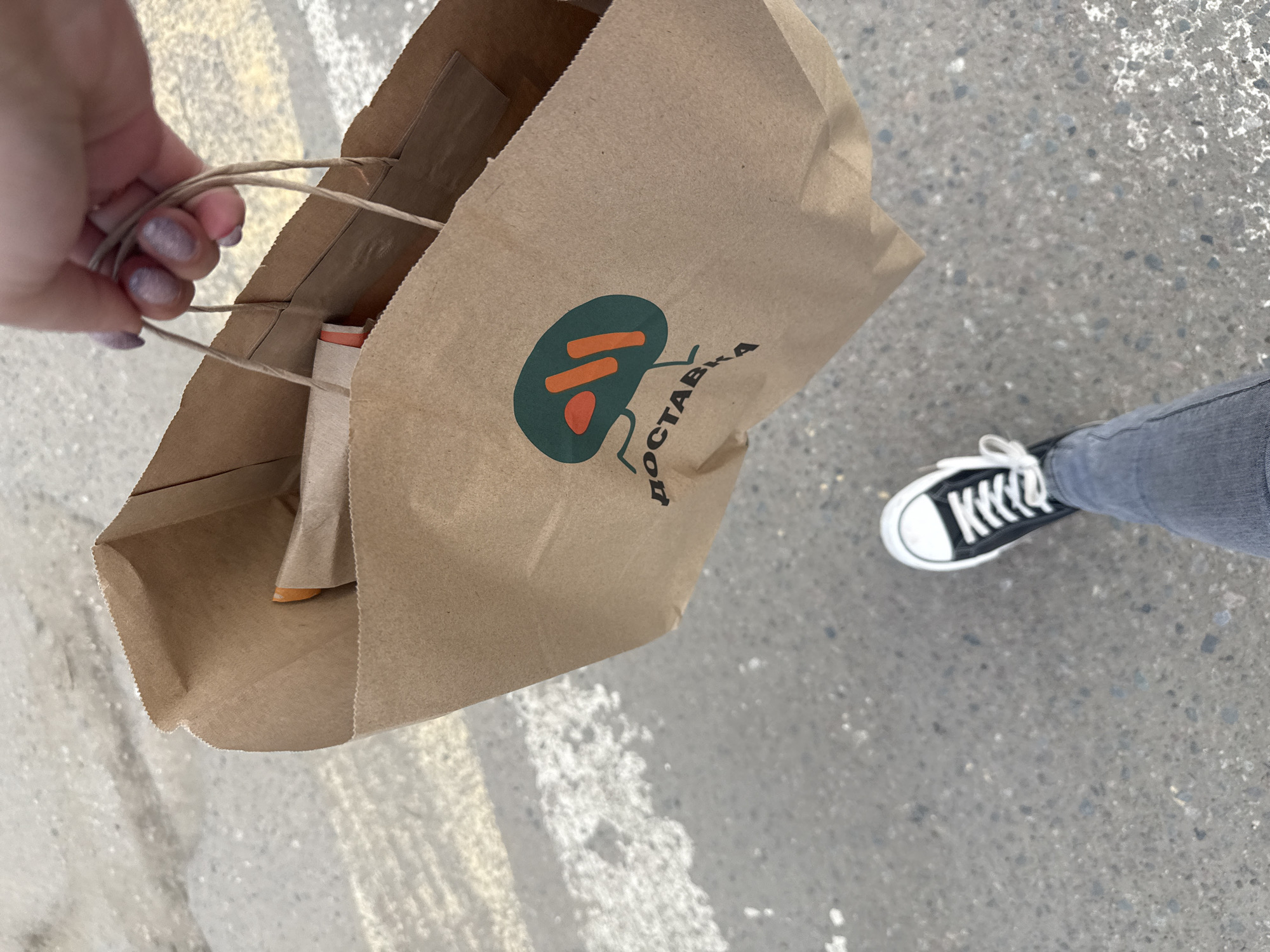 A top-down shot shows a person holding a brown paper bag with the logo of a restaurant or delivery service in what appears to be Dmitrov, Russia. The logo is green with two orange lines and a red dot, accompanied by the word "доставка," which means "delivery" in Russian. The bag seems to contain food, as an orange container is visible inside. The person is wearing gray jeans and black and white Converse-style sneakers. The environment is a gray concrete surface, likely a sidewalk or street. White and yellow stripes are painted on the surface, indicating a pedestrian crossing or some kind of lane marking. The hand holding the bag has light skin and sparkly nail polish.