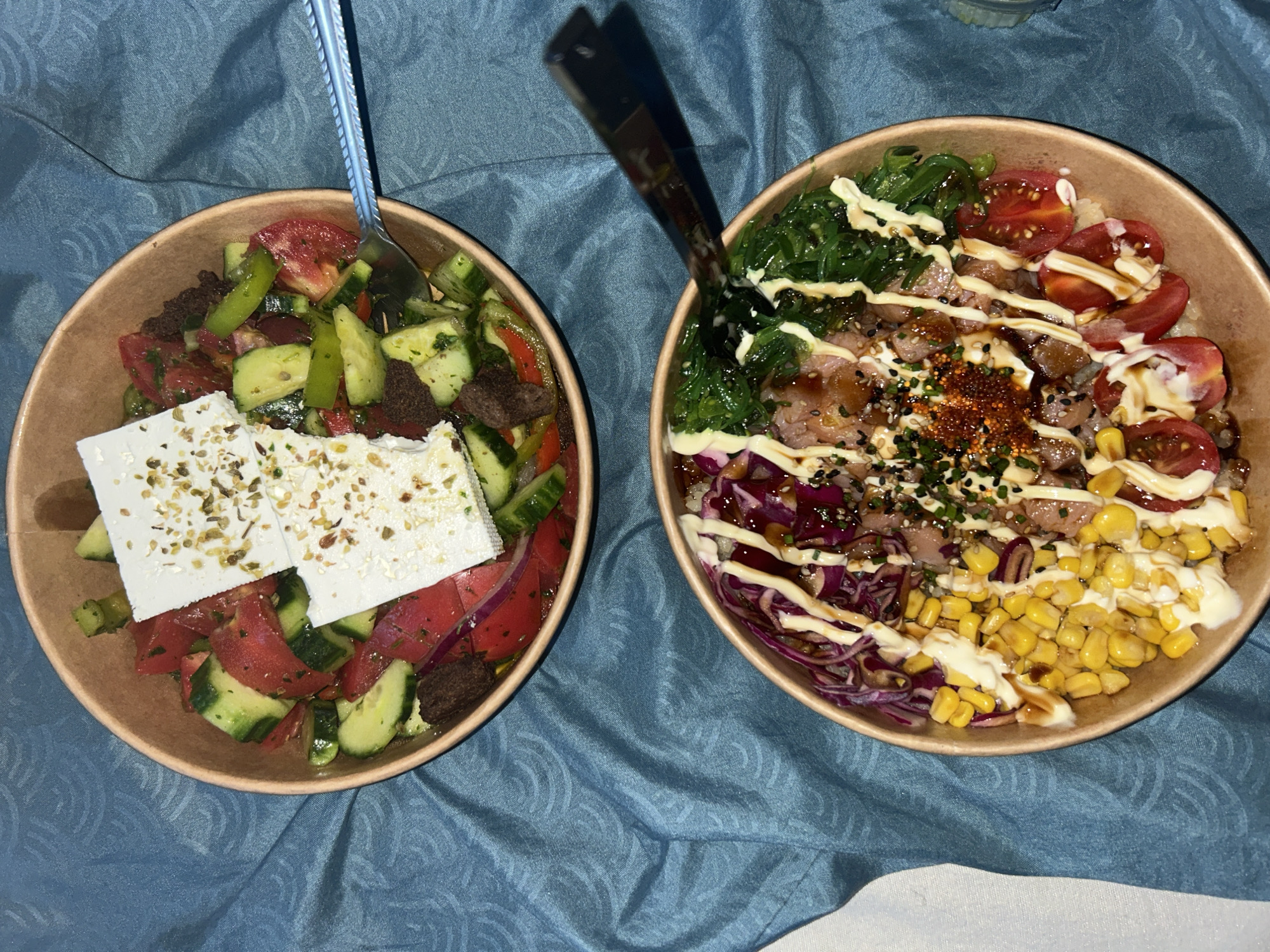 Two brown paper bowls of food are presented on a blue, textured fabric surface. The bowl on the left contains a Greek salad with chopped tomatoes, cucumbers, red onion, and chunks of dark bread. Two large pieces of feta cheese are placed on top, sprinkled with herbs. A silver fork is visible in the upper left of this bowl. The bowl on the right appears to be a poke bowl with rice at the bottom, topped with seaweed salad, cubed raw fish, cherry tomatoes, shredded red cabbage, and corn. Drizzled over the poke bowl are white sauce and a dark sauce, with a sprinkle of chili flakes and sesame seeds. Black chopsticks are resting on the edge of this bowl, with their tips in the food. The lighting suggests it is either daytime or indoors with artificial light. The food and presentation suggest a casual dining experience. No people are visible in the image. There is no visible text.