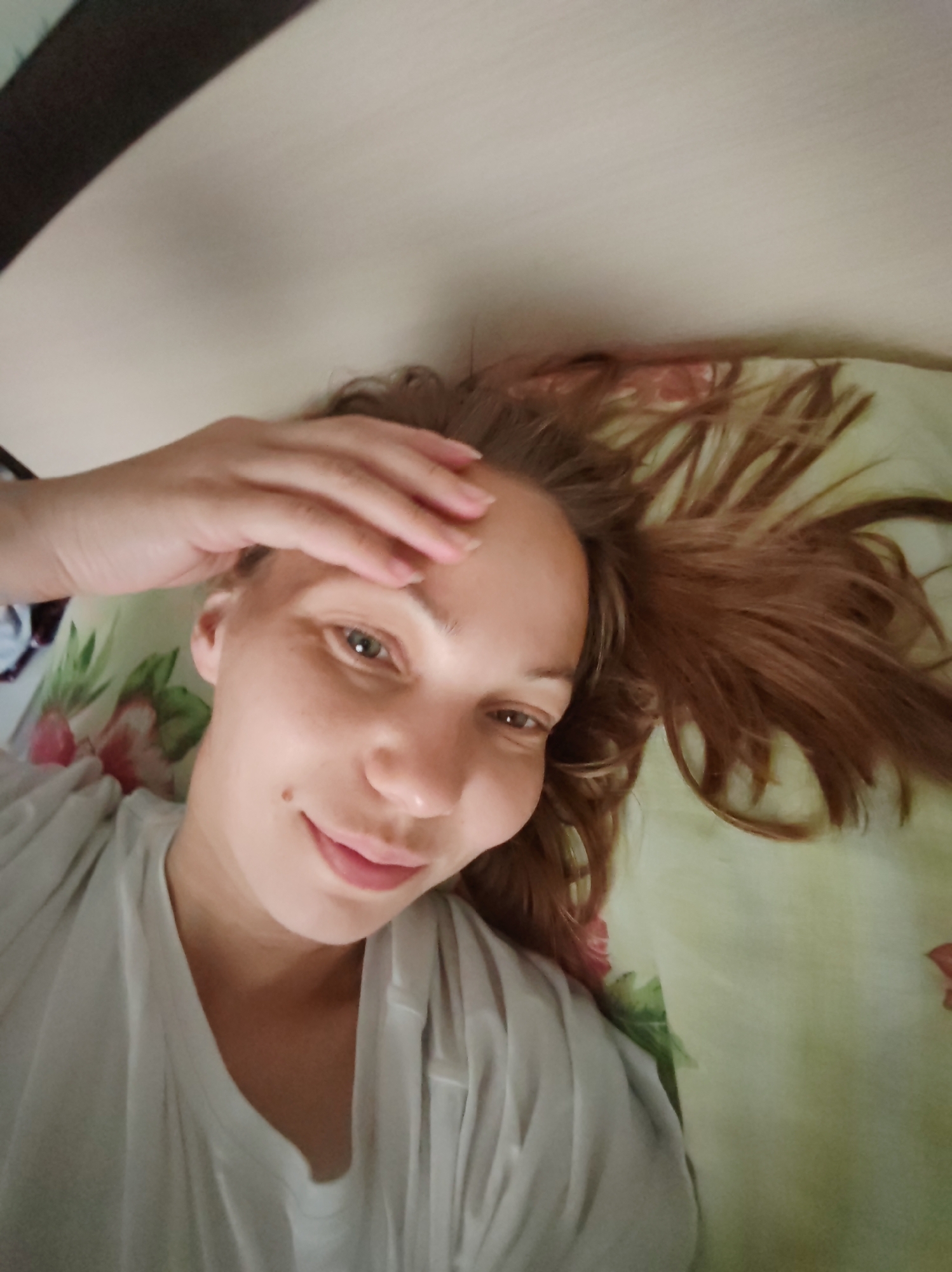 A young Caucasian woman with reddish-brown hair is lying on a bed, looking up at the camera. She is wearing a white t-shirt and has her left hand on her forehead. Her hair is spread out on a light green pillow with a floral pattern. The background includes a dark surface, possibly a headboard, and a portion of a wall or ceiling. The image appears to be a selfie taken indoors, likely in a bedroom. There is no indication of the specific location within Чухлэм, Russia.