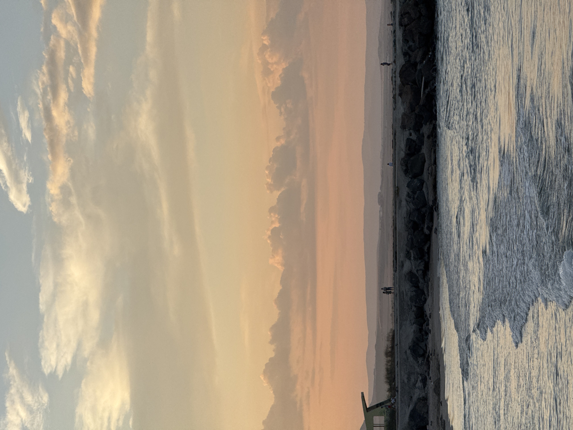 A serene, wide-angle shot captures the Pomorie coastline in Bulgaria during a sunset. The sky transitions from a pale blue at the top to soft hues of peach and yellow, adorned with wispy, illuminated clouds. Below, a distant, hazy mountain range forms the horizon. In the mid-ground, a man-made breakwater, constructed from large, dark rocks, juts out into the calm, rippling sea. Along the breakwater, a few silhouetted figures are visible, some walking, others standing and observing the view. On the far left, a modern, green-roofed structure with angled supports stands near the breakwater, possibly a shelter or part of a pier. The foreground is dominated by gentle waves lapping at the shore, reflecting the warm tones of the sky. The overall scene evokes a sense of peace and natural beauty.