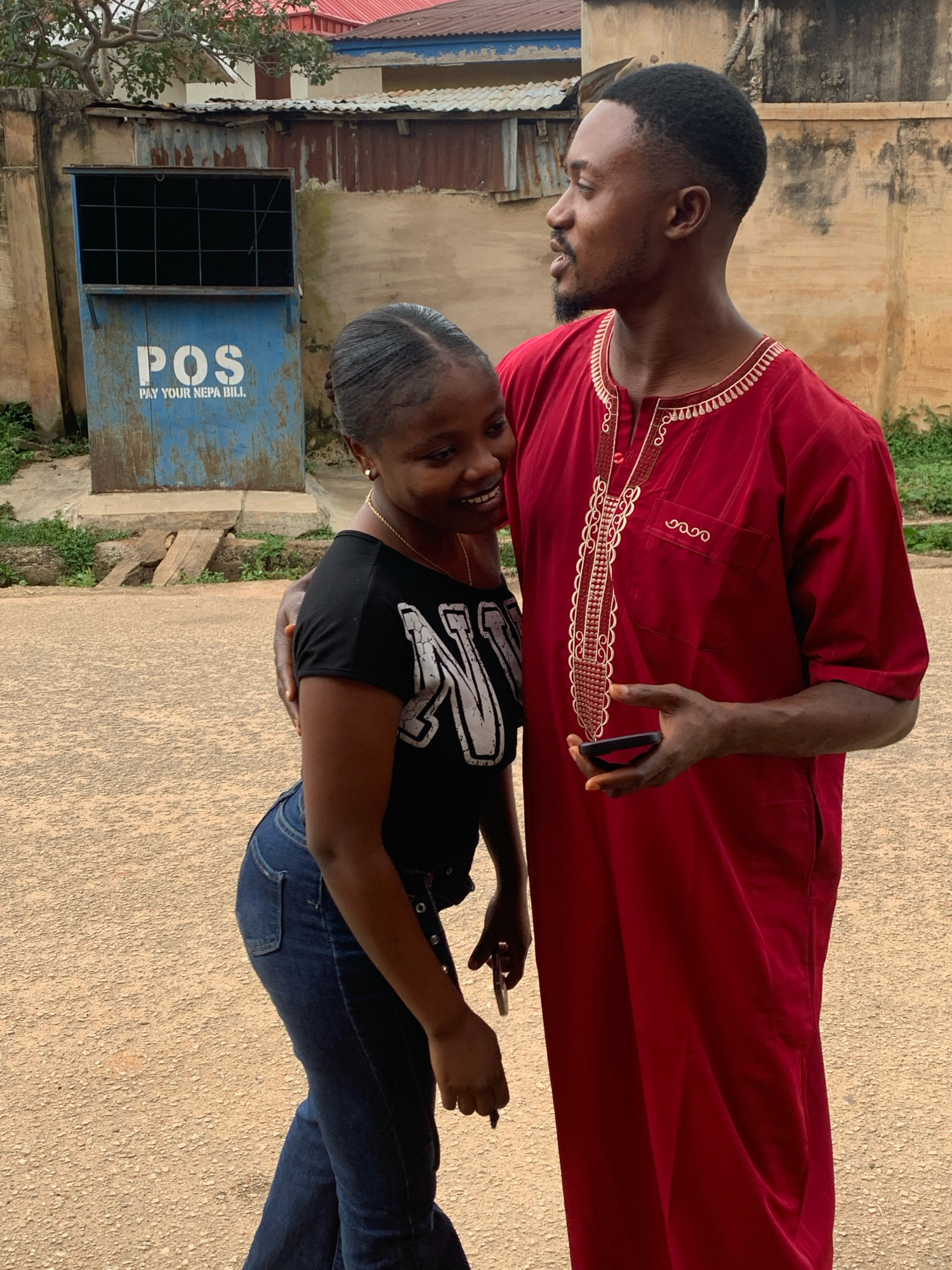 A young Black couple is posing for a photo in front of a weathered blue and tan POS (Pay Your Nepa Bill) kiosk, located in a street setting within Jos, Nigeria. The man, dressed in a red traditional African garment with white ornate embroidery, stands to the right, looking towards the woman. He has short hair and a goatee and is holding a mobile phone in his right hand. The woman, to the left, wears a black t-shirt with a white graphic and dark blue jeans. She is looking down and smiling, with her left arm around the man's waist and his right arm around her. Behind them, the POS kiosk is mounted on a concrete base. The background consists of a textured wall, a corrugated metal roof, and some sparse greenery. The ground is an asphalt or paved surface.