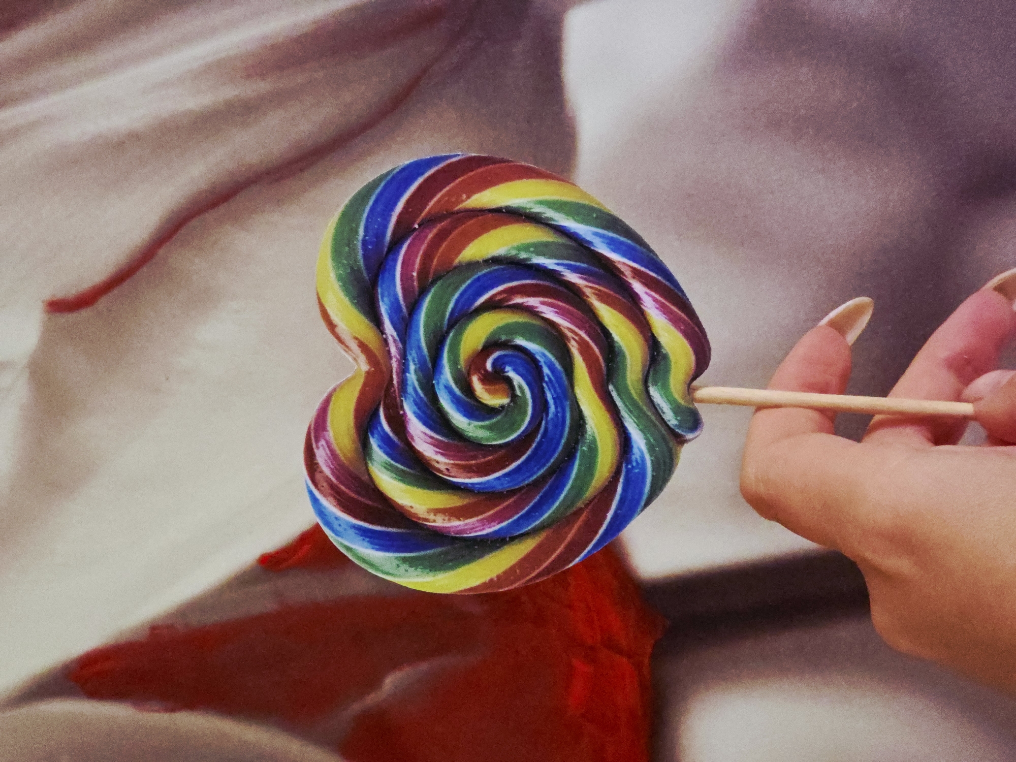 A person with long, manicured nails is holding a multi-colored, spiral lollipop on a wooden stick. The lollipop appears to be heart-shaped. The background features a blurred, abstract pattern of beige, white, and red. The location and city are unknown.