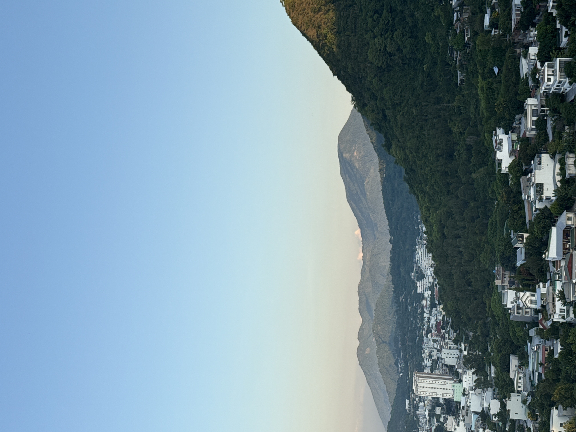 This is an aerial shot of a coastal city in Nha Trang, Vietnam, with mountains in the background. The city is densely populated with a mix of low-rise buildings and a few taller structures, including a prominent white high-rise building. Lush green trees and vegetation cover the hillsides, encroaching into the urban areas. The sky is clear and a light blue, with the sun casting a warm glow on the landscape, suggesting it is either early morning or late afternoon. The image captures a serene view of the city nestled between the mountains and the sea, a common characteristic of Nha Trang's geography. No people or specific actions are visible in the image.